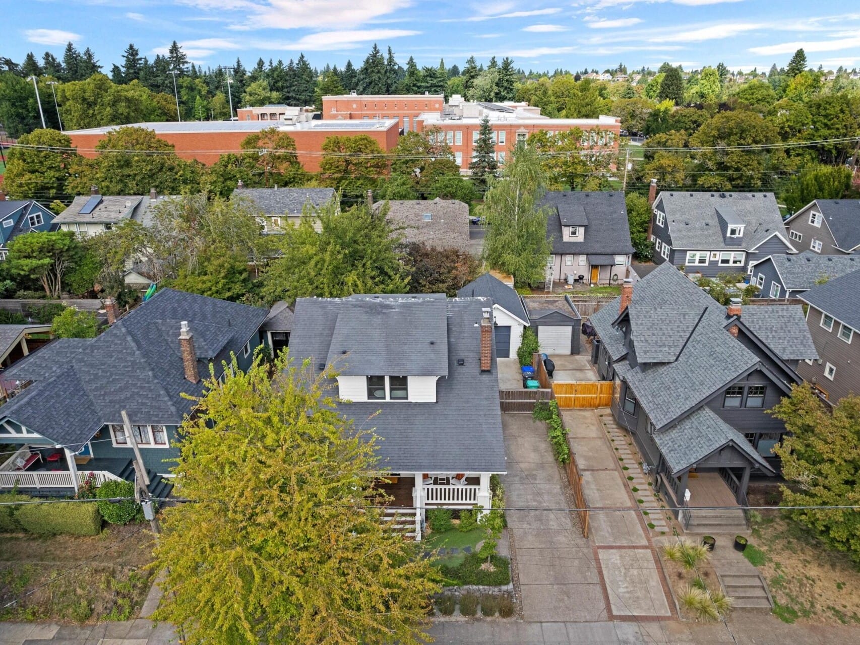 Aerial view of a Portland, Oregon residential neighborhood with several houses featuring grey and darker roofs, surrounded by trees. In the background, there's a large red-brick building and a sports field. The sky is partly cloudy.