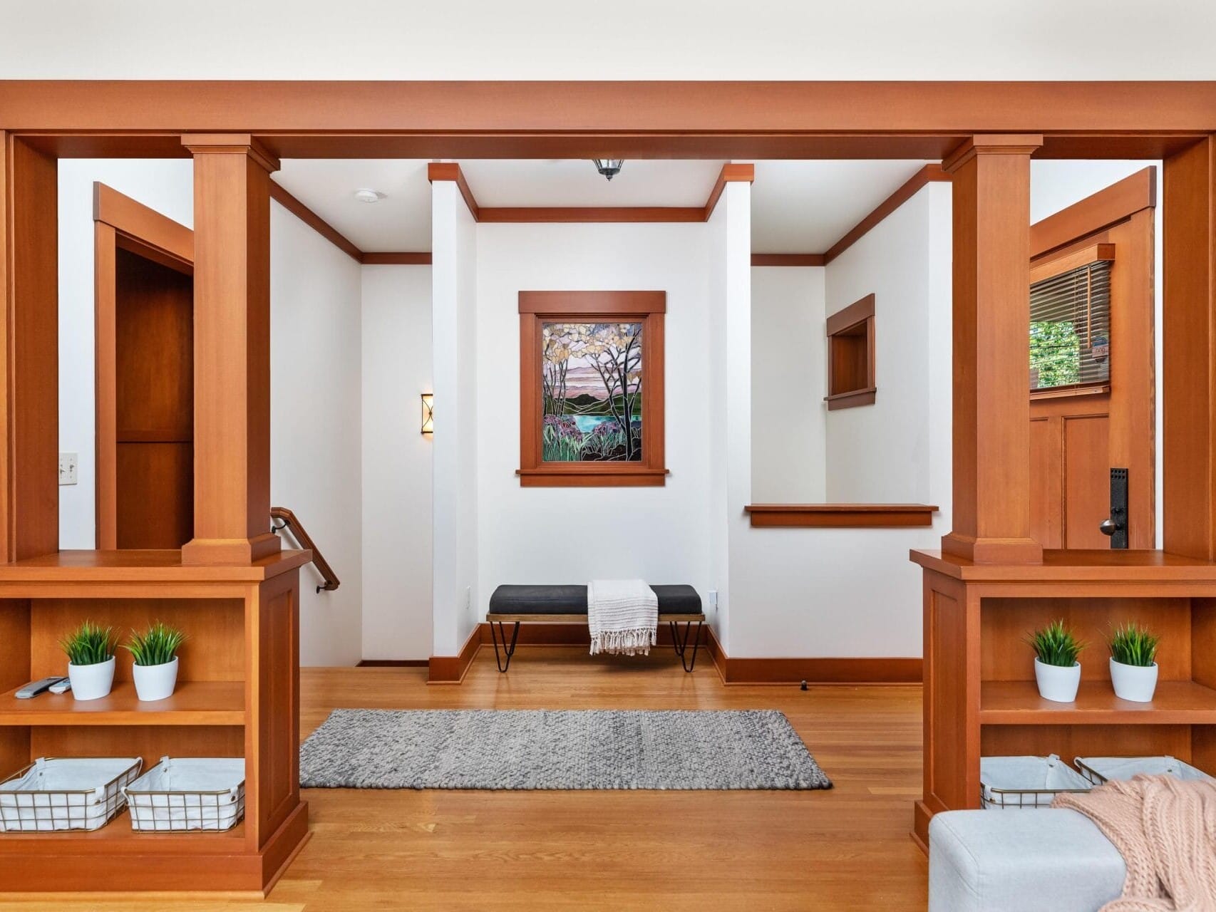 A modern Portland entryway features wooden columns and built-in shelving. Two small plants and baskets adorn the shelves. A grey bench with a blanket rests beneath a window, while a textured rug complements the wooden floor, creating an inviting atmosphere typical of Oregon's design sensibilities.