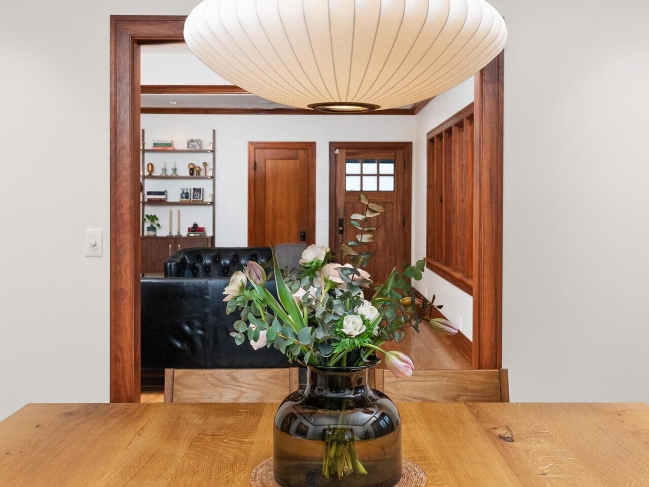 A modern dining area with a wooden table and a vase of flowers is elegantly lit by a globe-shaped pendant light. In the background, glimpse a cozy living room, perfect for showcasing to clients by your top Portland real estate agent.