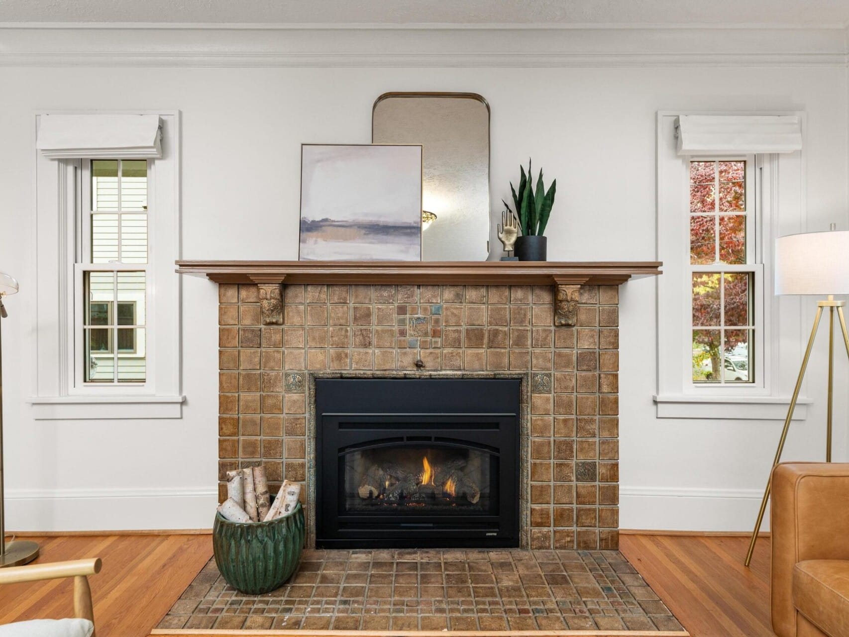 Cozy living room with a tile fireplace, framed by two windows with white blinds. A painting, a mirror, and a plant are on the mantel. A basket with logs is beside the fireplace. A leather chair and modern floor lamp are on the right.