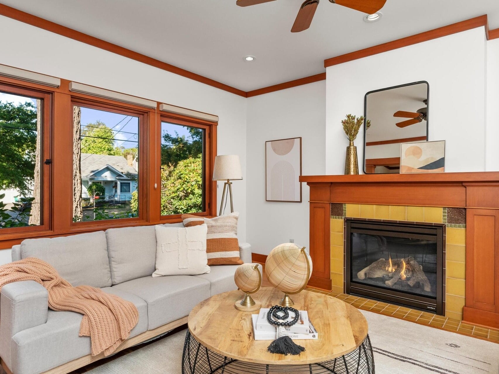 A cozy living room in Portland, Oregon, features a beige sofa with an orange throw blanket and pillows near large windows. A wooden coffee table with decorative items sits in front of a lit fireplace, while minimalist art and a large mirror adorn the walls above the mantel.