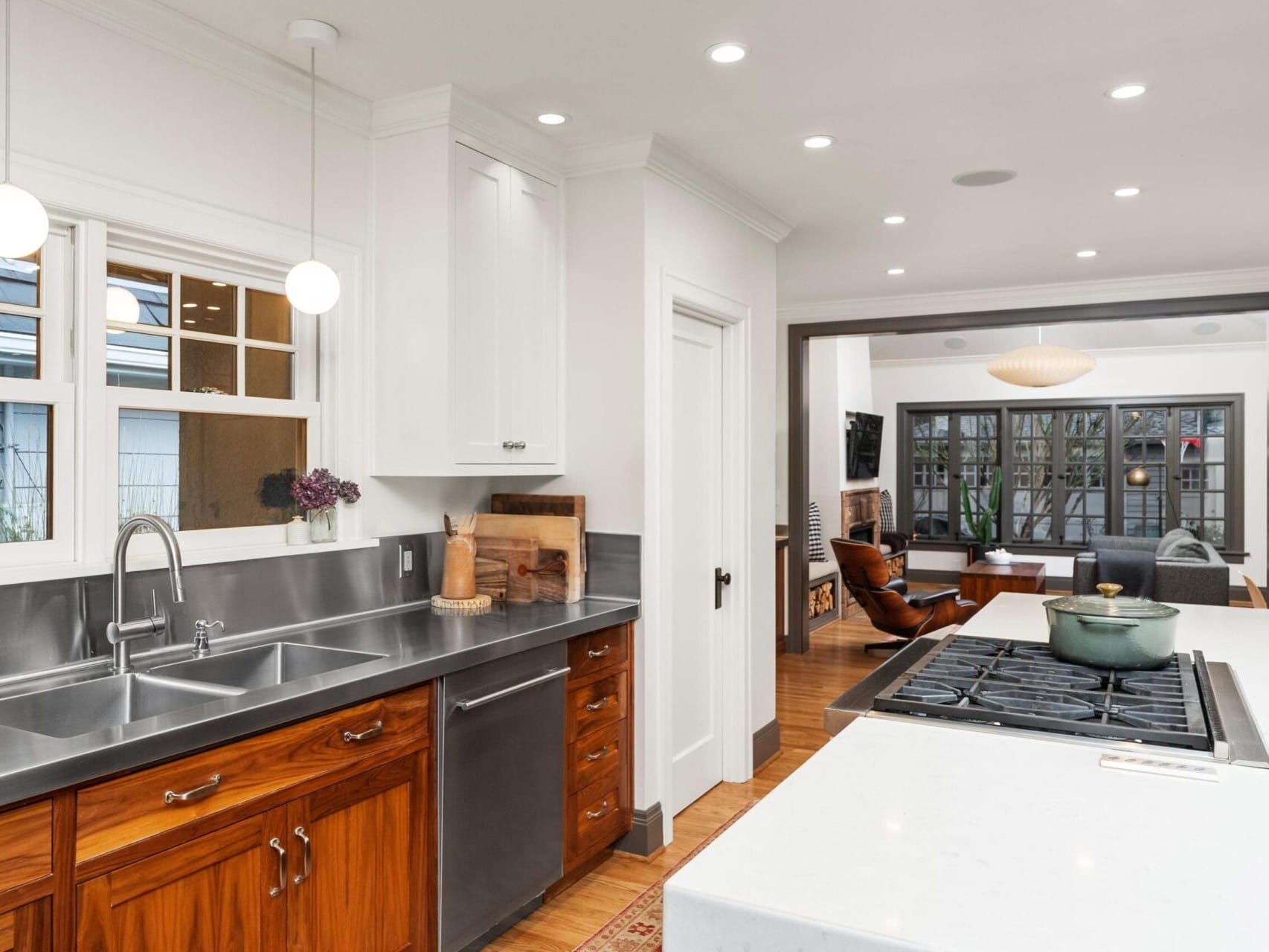 A modern kitchen with wooden cabinets and a stainless steel countertop. There's a large island with a stove and pot, illuminated by pendant lights, offering an open view to a cozy living area. Discover this gem through your Portland Oregon Real Estate expert for the perfect home experience.