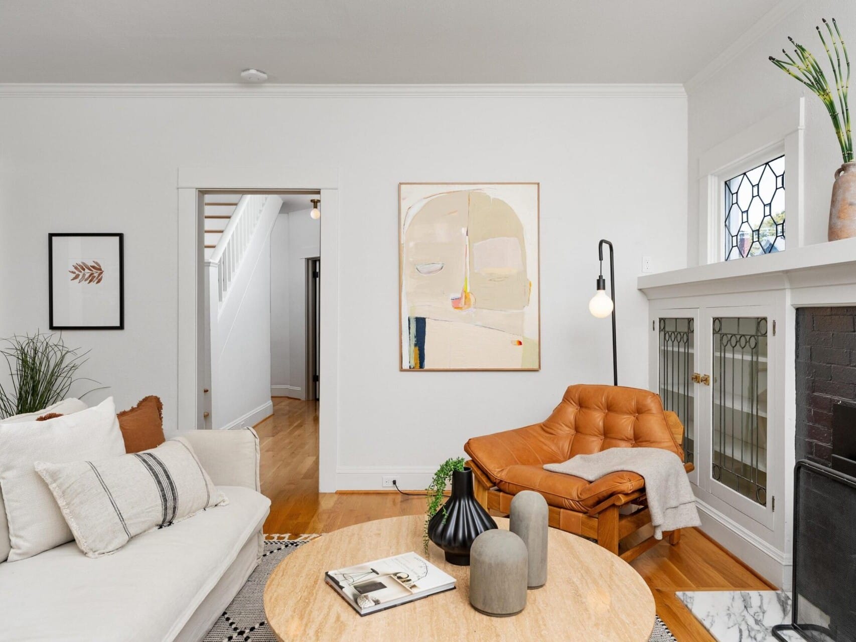 A cozy Portland, Oregon living room features a white sofa, an orange chair, and a round wooden coffee table. Art pieces and vases with plants decorate the space. A modern abstract painting hangs on the wall above the sofa, and there's a fireplace on the right.