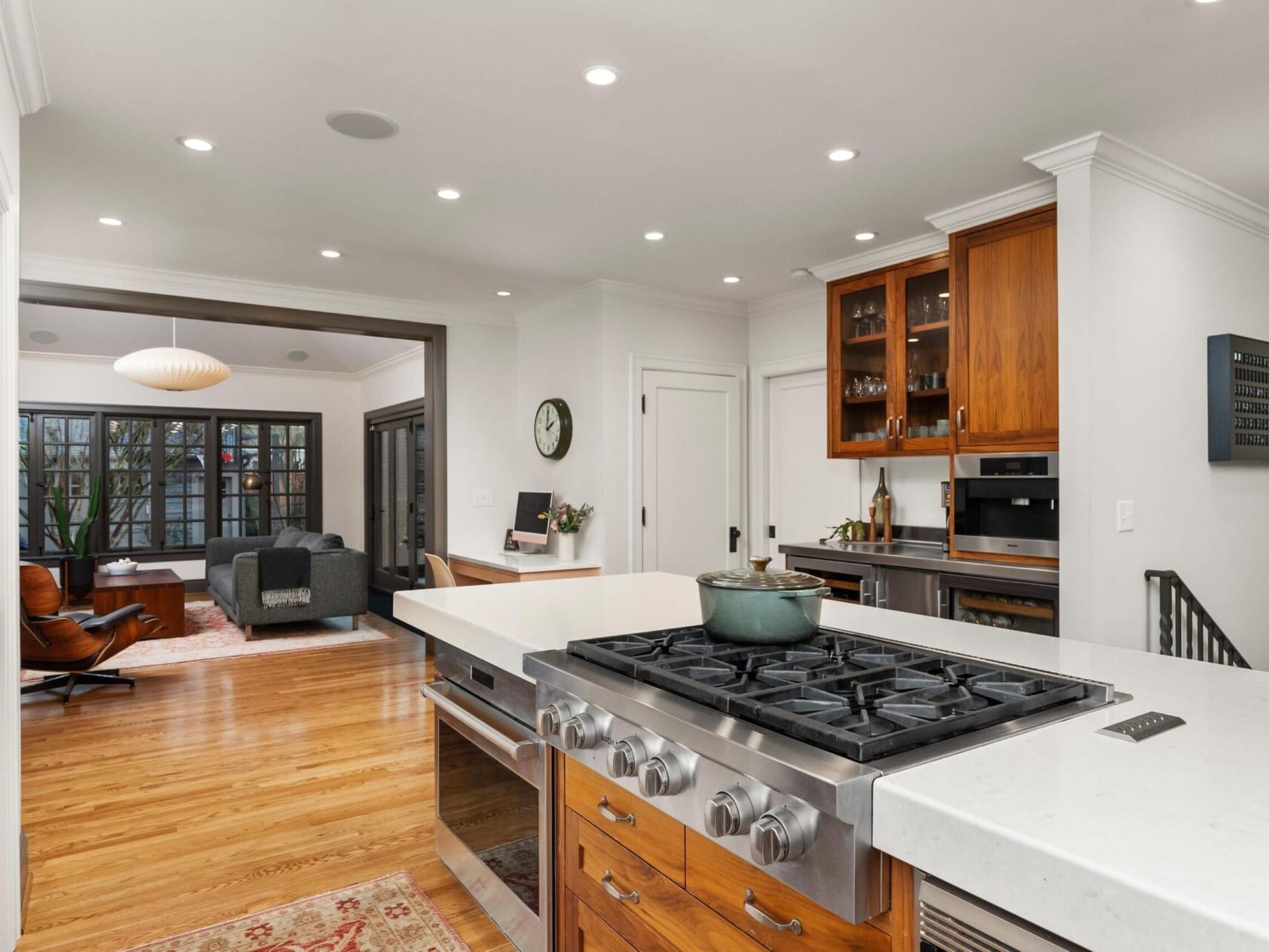 Modern kitchen with a large white island, built-in gas stove, and wooden cabinets. The kitchen opens to a cozy living room with a dark sofa and large windows. Hardwood floors and recessed lighting add warmth and brightness to the space—a gem in Portland Oregon real estate.