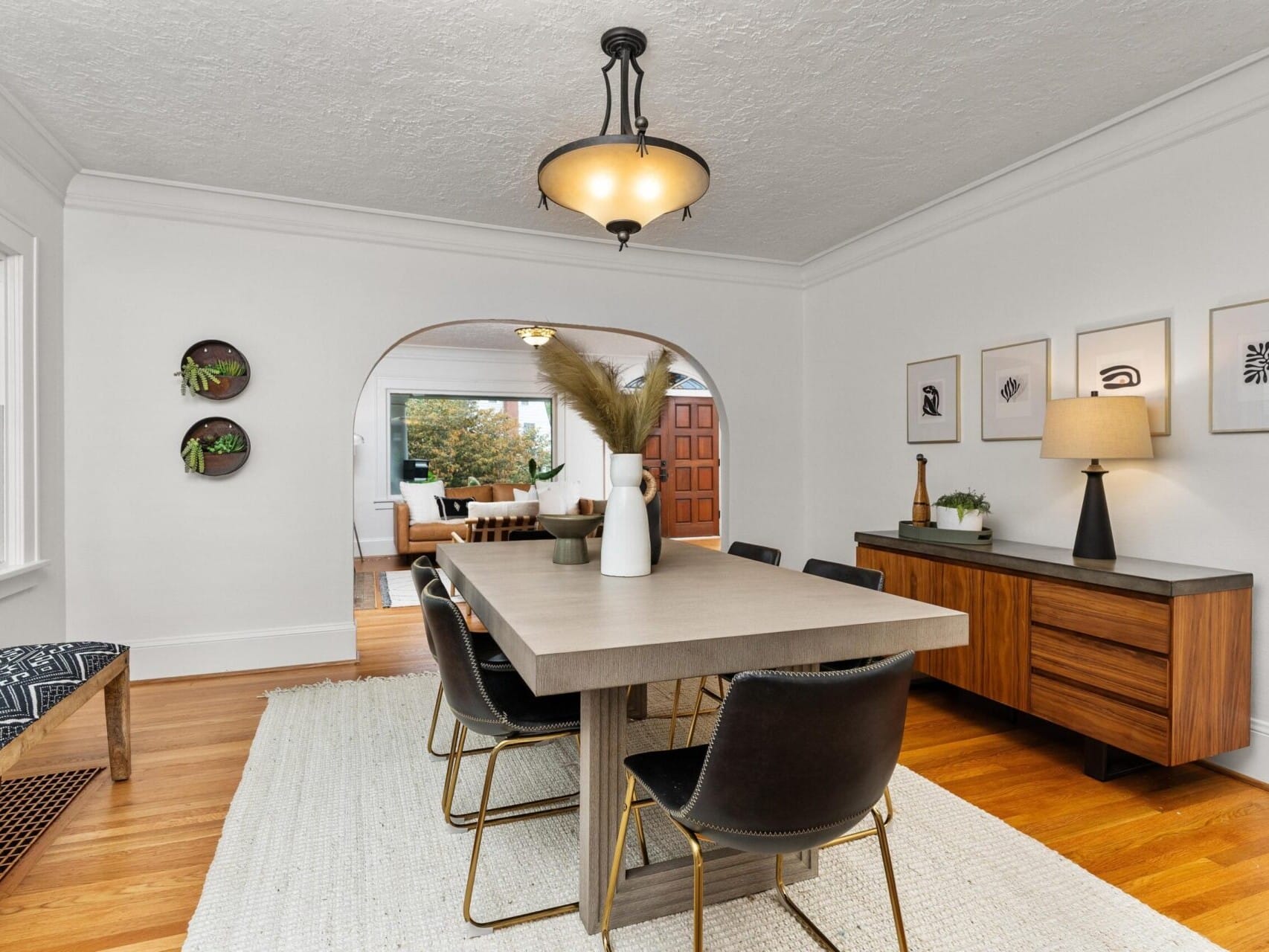 A modern dining room with a wooden table and six black chairs on a white rug. The walls are decorated with small plants and framed artwork. A chandelier hangs above, and the room opens into a living space with a couch visible in the background.