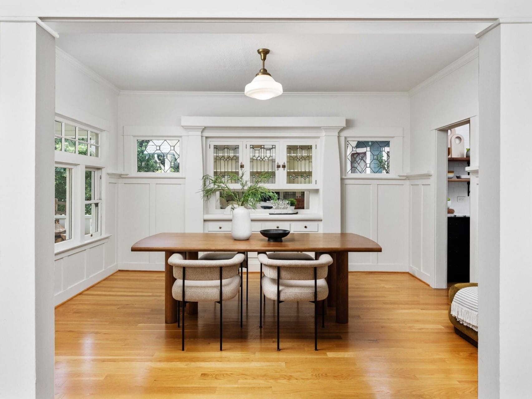 A bright dining room in Portland, Oregon, with white walls and a wooden floor features a large wooden table with four upholstered chairs. Decorative cabinets and windows with stained glass accents adorn the back wall. A single pendant light hangs above the table.