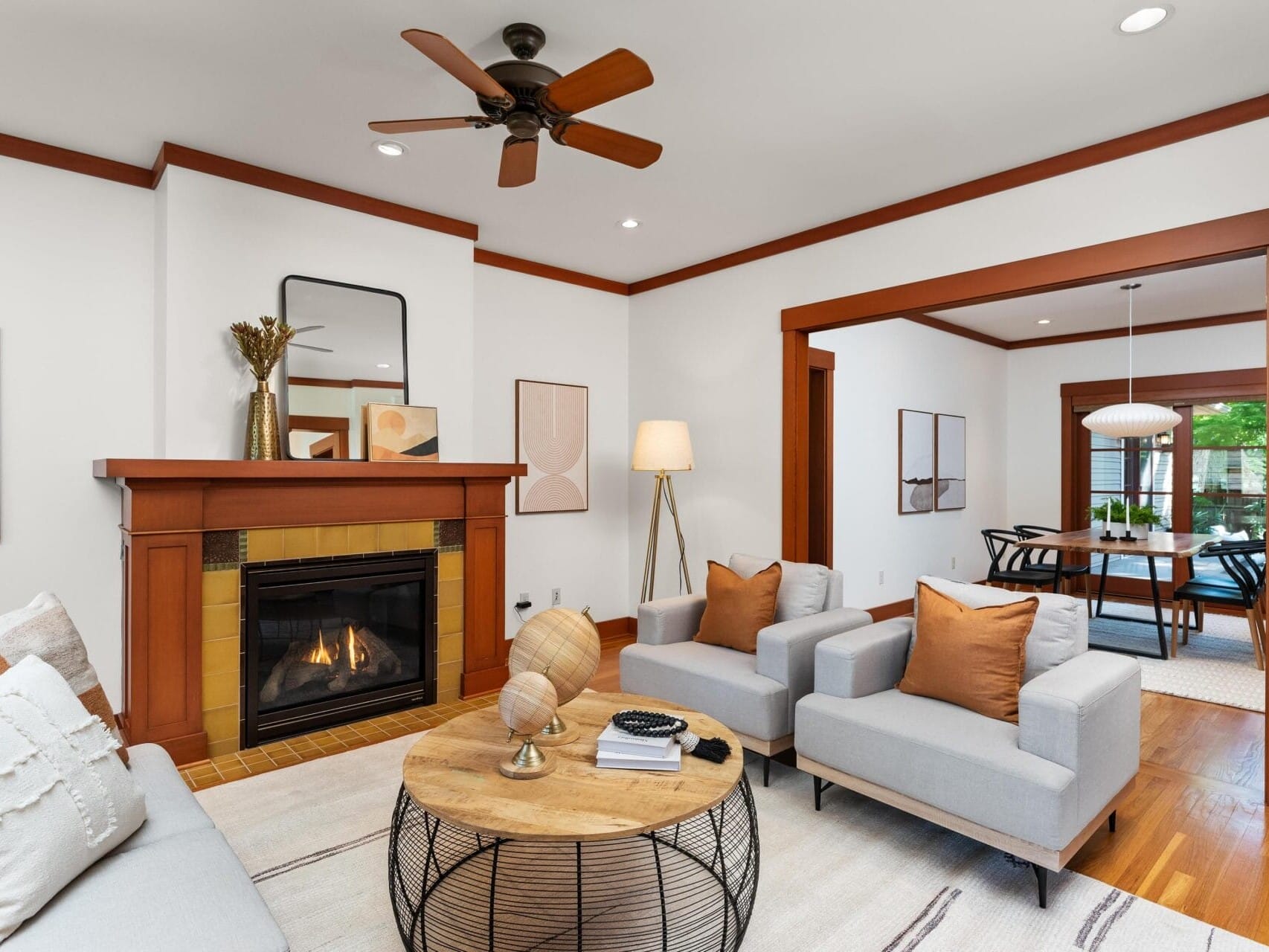 A cozy living room with a warm color scheme reminiscent of Portland, Oregon, features two light gray armchairs with brown cushions, a wooden coffee table, and a fireplace. A ceiling fan is above, and a dining area is visible through a wooden-framed doorway.