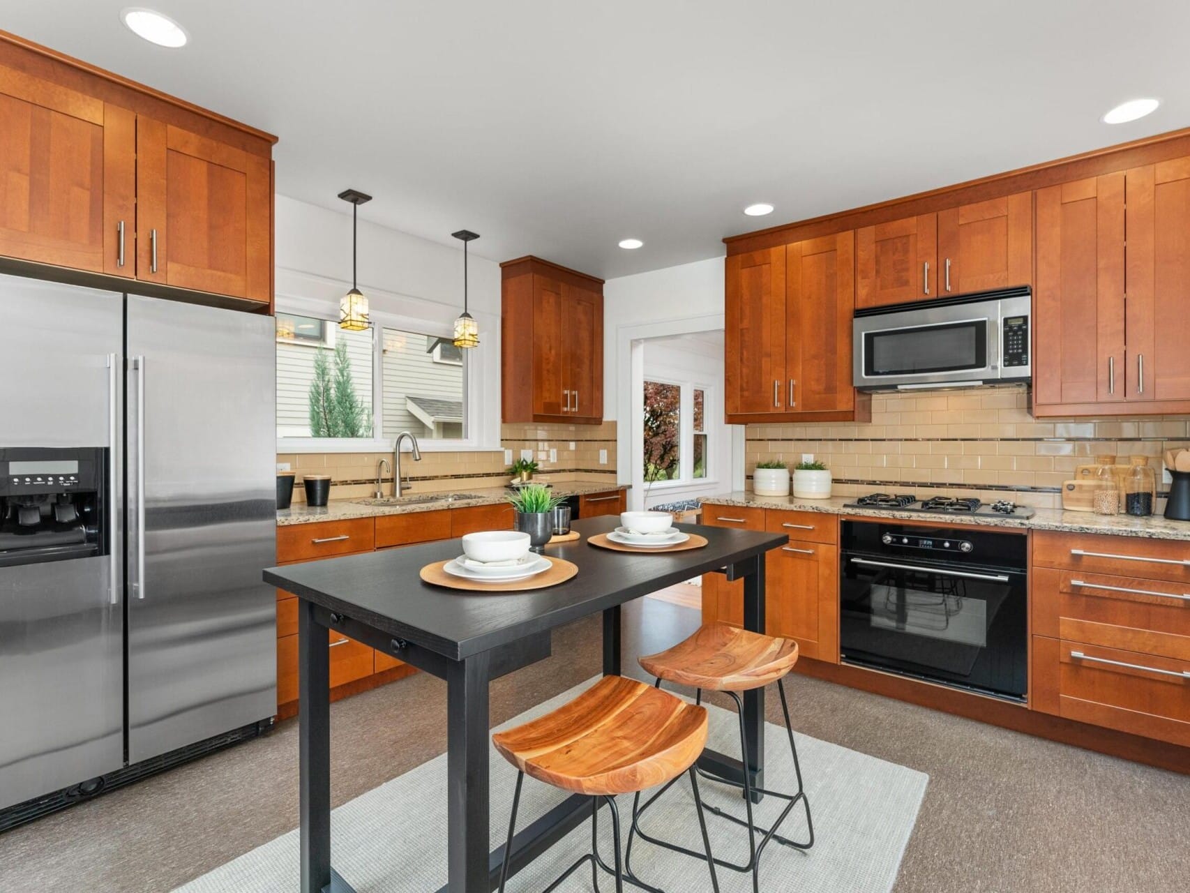 A modern kitchen with wooden cabinets and stainless steel appliances, including a refrigerator and built-in oven. A black island with two wooden stools is centered, and pendant lights hang above. The room is well-lit with natural and recessed lighting.