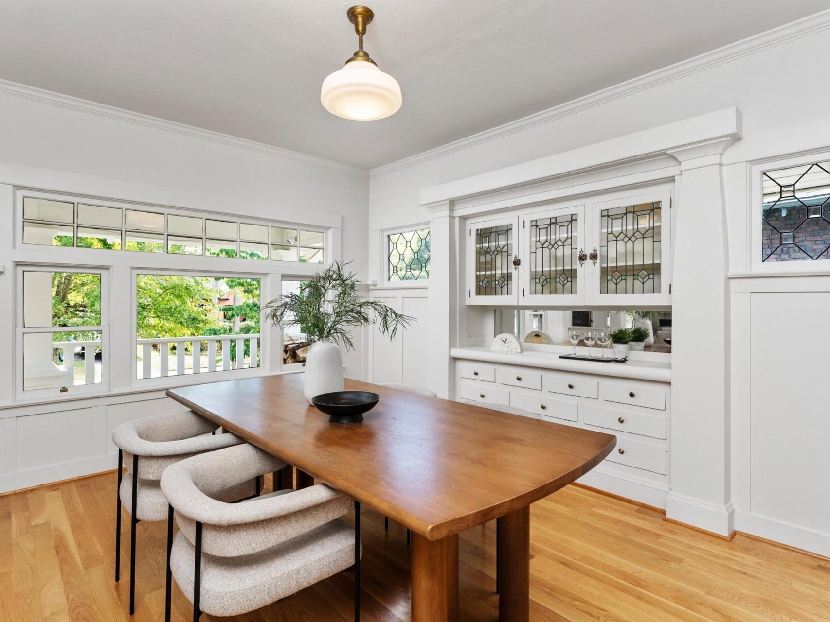 A bright dining room in Portland, Oregon features a wooden table surrounded by light gray chairs. A white built-in cabinet with glass doors and decorative displays graces the right wall. Large windows flood the space with natural light, offering a serene view of the lush greenery outside.