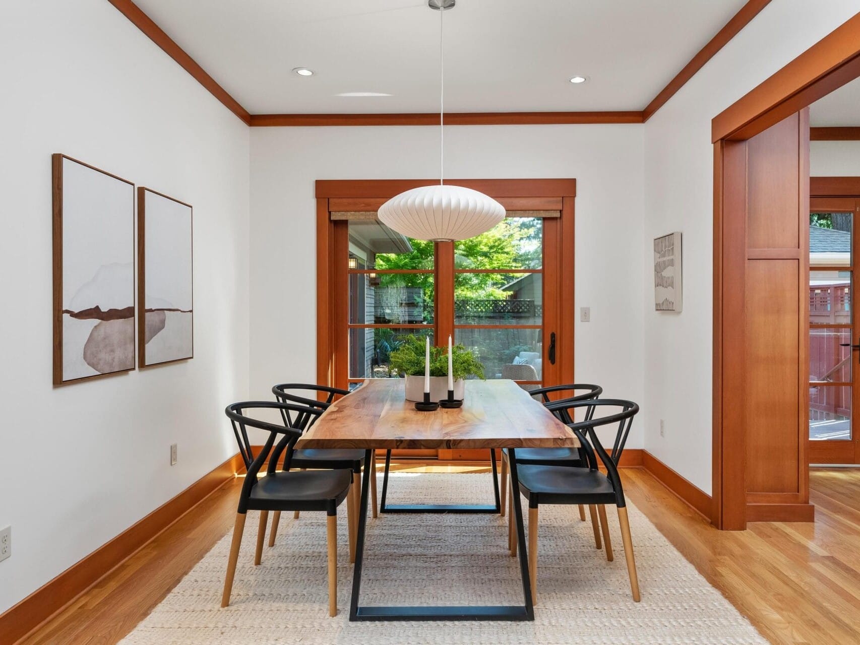 A modern Portland Oregon dining room features a wooden table with six black chairs on a light rug. A large white pendant light hangs above, while two abstract paintings adorn the white walls, and a glass door reveals lush greenery outside.