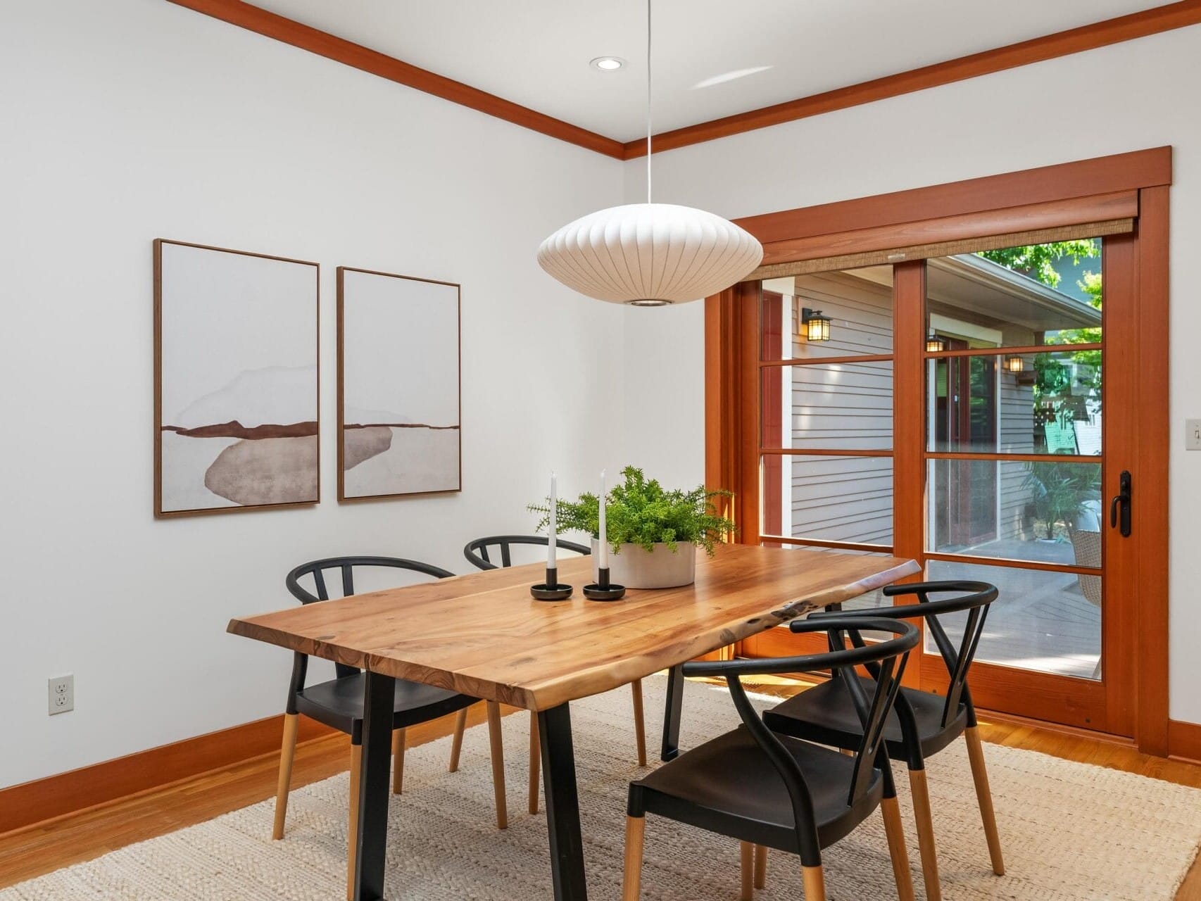 A modern dining room in Portland, Oregon, features a wooden table surrounded by black chairs. Above the table hangs a round, white pendant light. Two abstract paintings adorn the wall, while large sliding glass doors open to a scenic view outside.