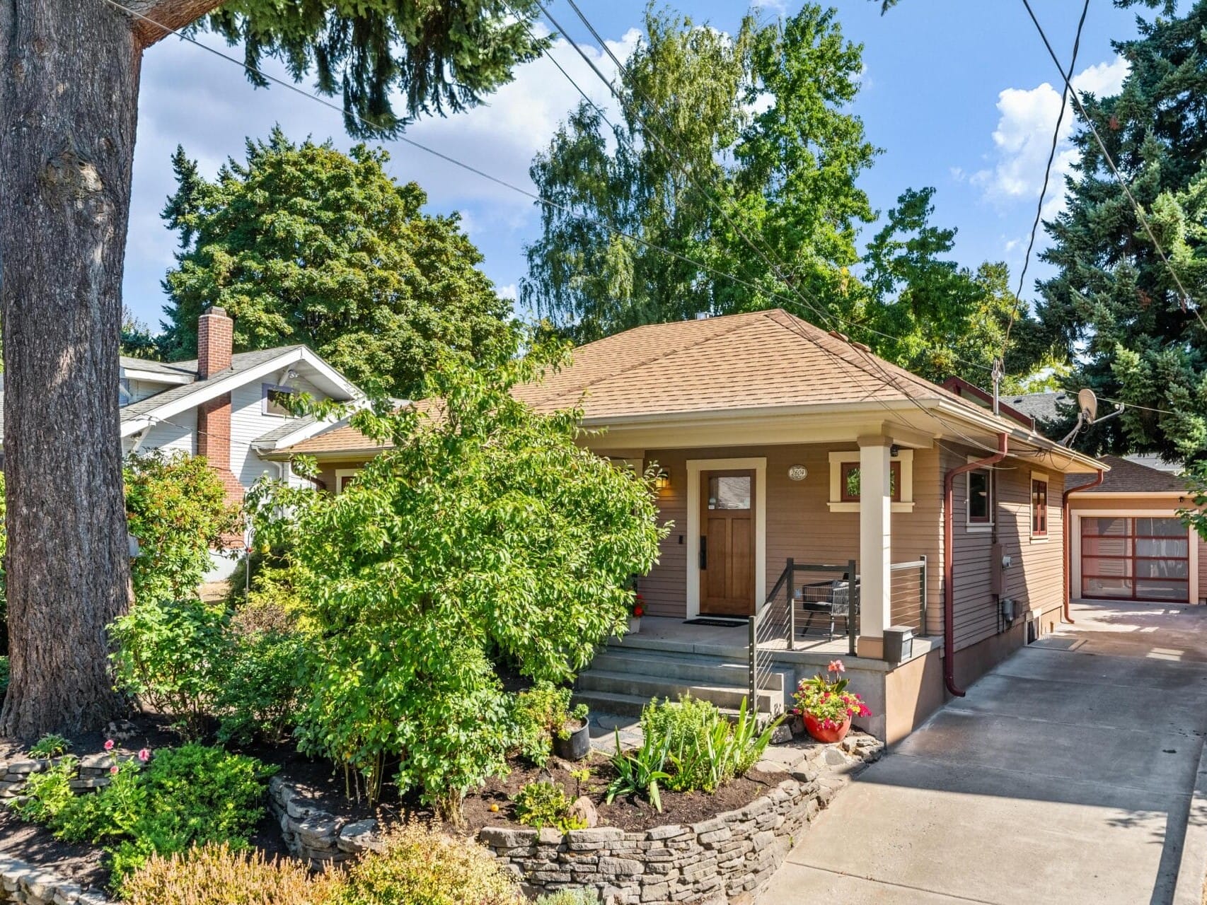 A charming single-story house in Portland, Oregon, features a beige exterior and covered porch, embraced by lush greenery and trees. A stone-bordered garden graces the front, while the driveway winds its way to a detached garage tucked in the back.