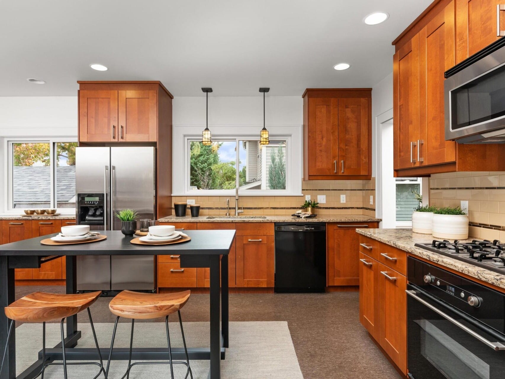 A modern kitchen with wooden cabinets, stainless steel appliances, and a central island with two wooden stools. The room is well-lit, featuring pendant lights and a window with a view of greenery outside.