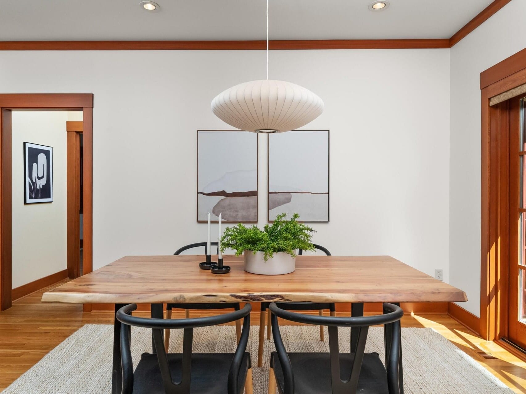 A modern dining room in Portland, Oregon, features a wooden table with six black chairs. A potted plant and two candle holders accentuate the table. A large pendant light hangs above, with abstract artwork on the wall. Natural light filters through glass doors, enhancing the serene setting.