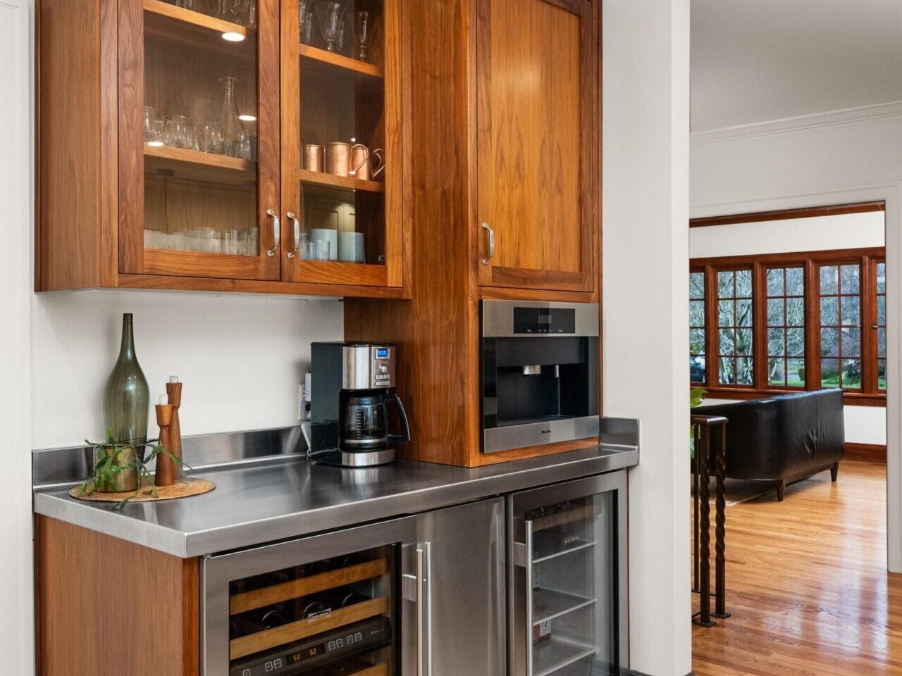 A modern kitchen alcove in Portland Oregon real estate, featuring wooden cabinetry and stainless steel appliances, including a wine fridge and coffee maker. Glass-paneled cabinets display glassware above a sleek countertop, while hardwood flooring complements the warm wood tones.