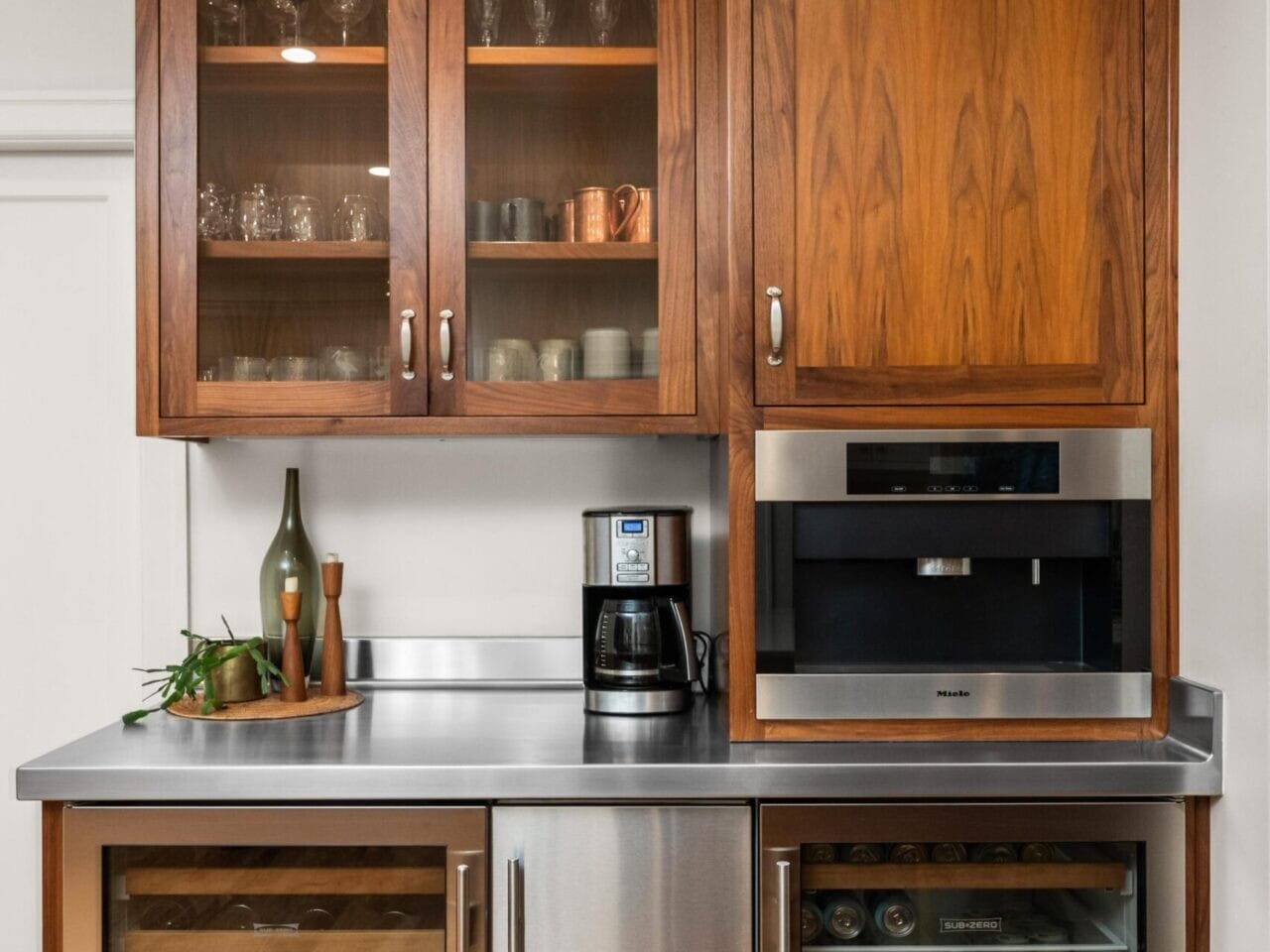 A home kitchen, ideal for any Portland Oregon real estate listing, features wooden cabinets with glass doors showcasing glassware, a coffee maker, and a built-in microwave. Below, a dual-section refrigerator stores wine bottles on the left and beverages on the right. The countertop is stainless steel.