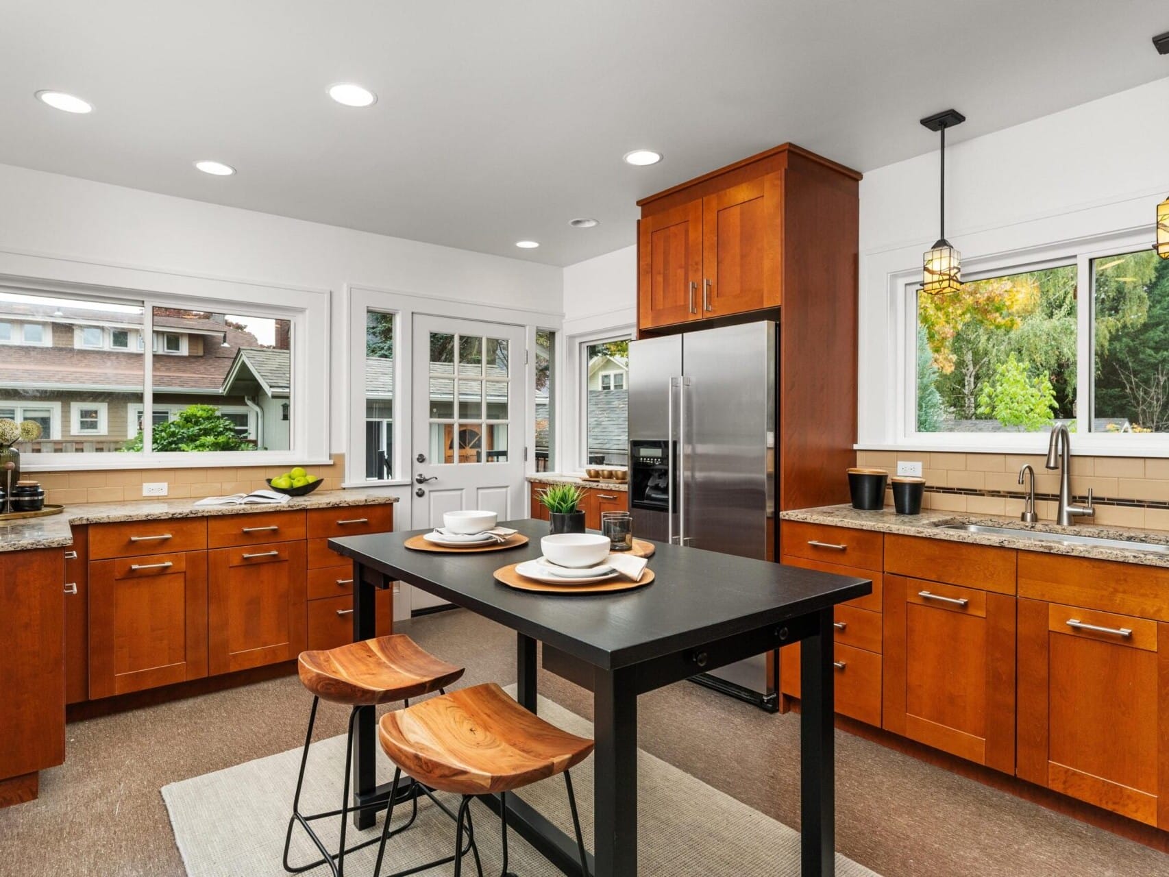 A modern kitchen with wooden cabinets, stainless steel appliances, and a central island with two wooden stools. The room is well-lit with pendant lights and natural light from large windows, offering a view of greenery outside.