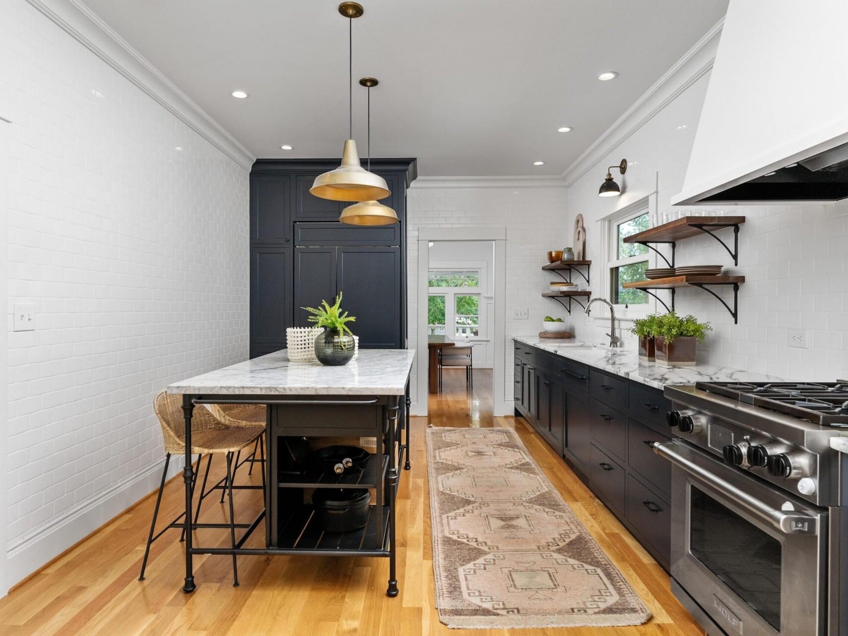 A modern kitchen in Portland, Oregon, with white walls and sleek black cabinetry. It features a central island with stools, pendant lights, and a stove with a white hood. Open shelves hold decorative plants, while a patterned rug stretches across the light wood floor.