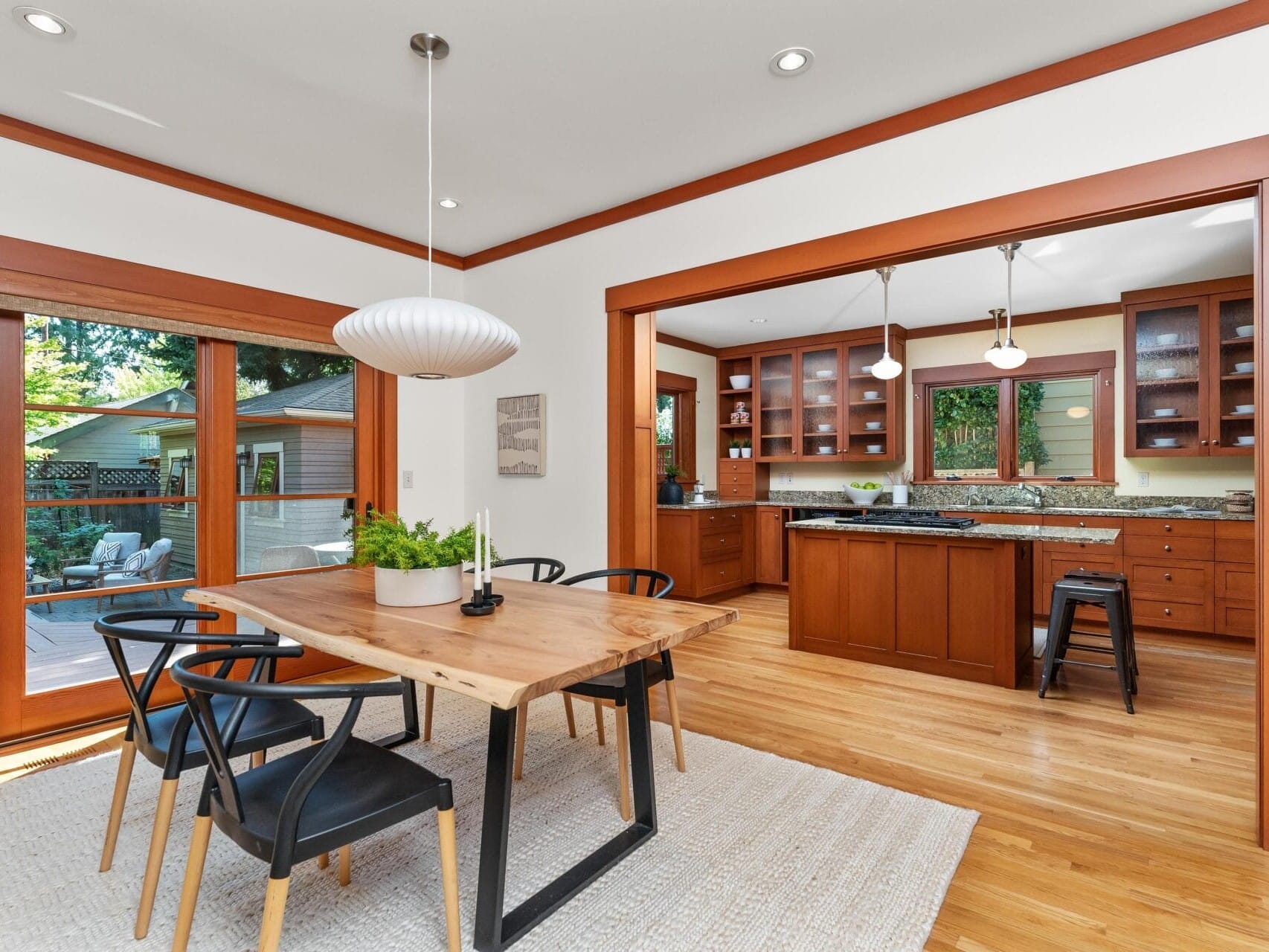 A dining area with a wooden table and black chairs flows into an open kitchen featuring wooden cabinetry and bar stools, embodying Portland, Oregon charm. Hardwood floors, pendant lighting, and a large window door open to a garden.