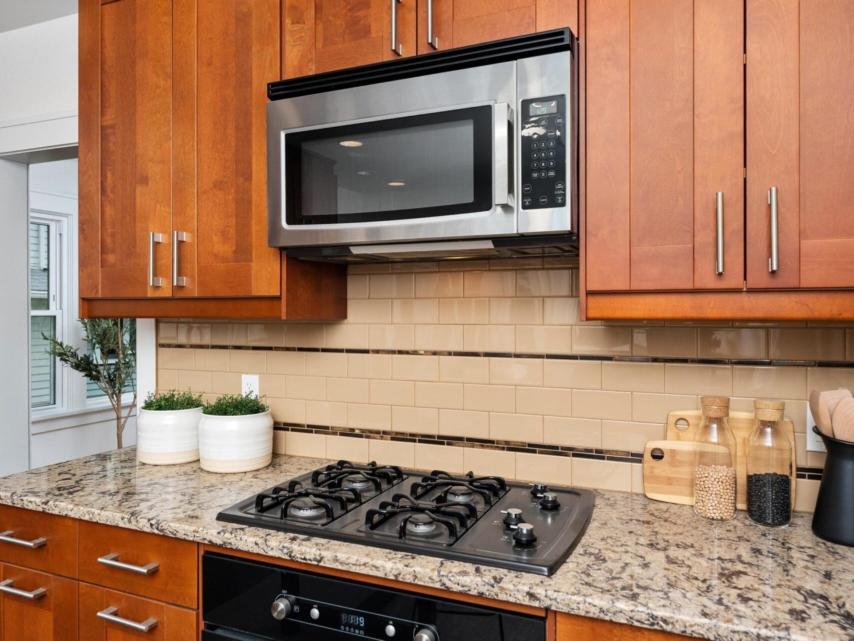 Modern kitchen with wooden cabinets, a granite countertop, and a gas stovetop. A stainless steel microwave is mounted above the stove. There are plants and jars with ingredients on the counter, and beige tiles as a backsplash.