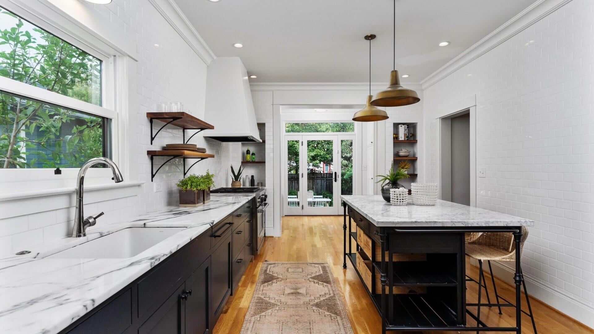 A modern kitchen in Portland, Oregon, boasts white walls and a wooden floor, featuring a long marble-topped island with pendant lights above. Open shelves hold plants and kitchenware, while large windows and a glass door offer views of the lush garden outside.