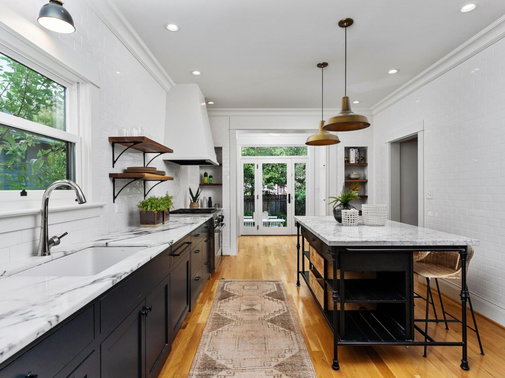 A modern kitchen in Portland, Oregon, features white walls, a marble countertop, and black cabinets. Two pendant lights hang above a central island with high stools. Natural light flows through a window and glass doors, revealing lush greenery outside.