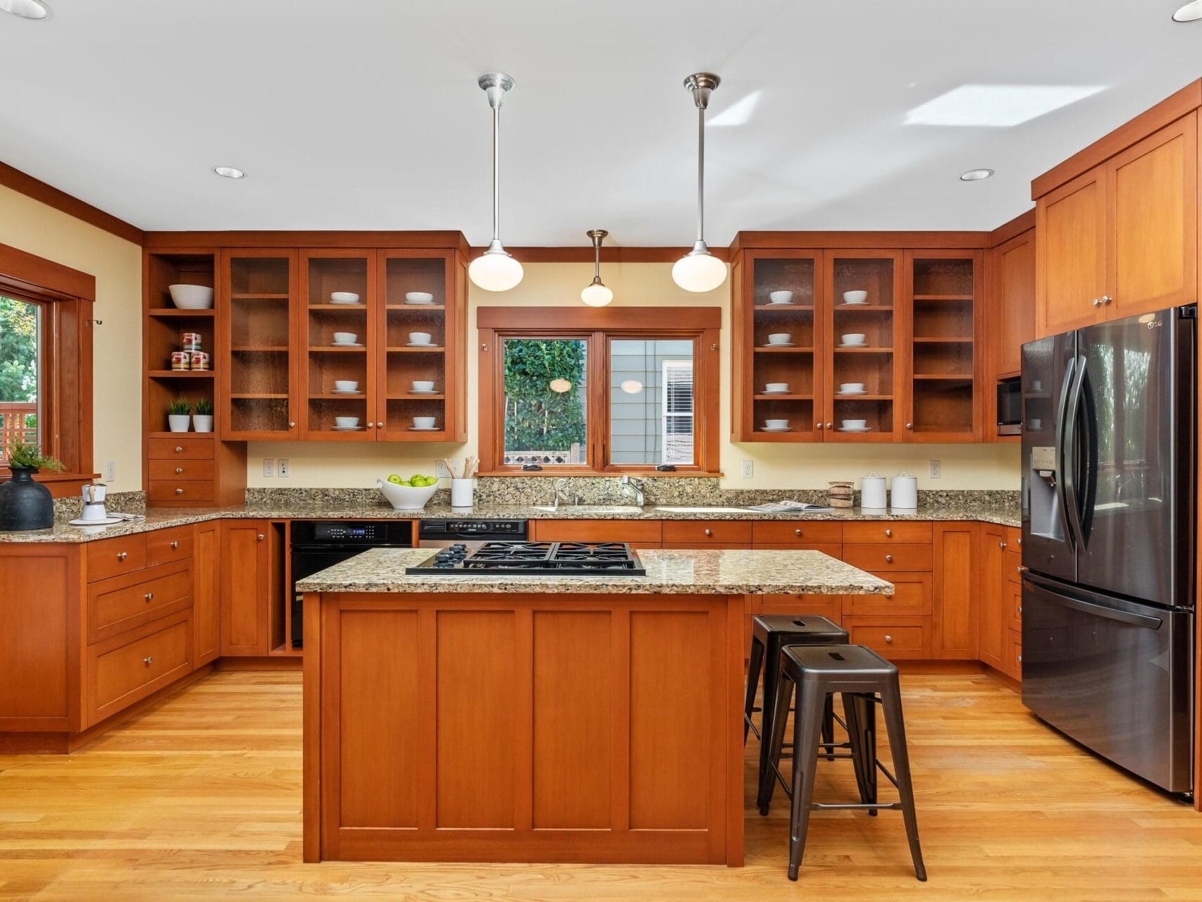 A modern kitchen in Portland, Oregon features wooden cabinets, granite countertops, and a center island with a stovetop. Two black stools sit at the island. Stainless steel appliances, including a refrigerator, blend seamlessly as natural light floods through a large window.