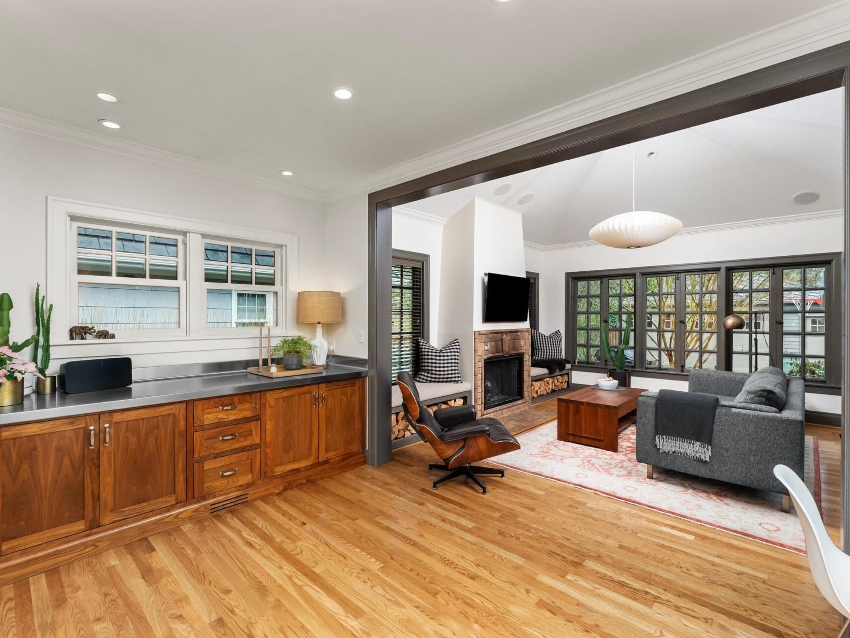 A modern living room with wooden flooring features a gray sofa, armchair, fireplace, and wall-mounted TV. Large windows let in natural light. A kitchen area with wooden cabinets and countertop is partially visible on the left, perfect for those exploring Portland Oregon real estate.