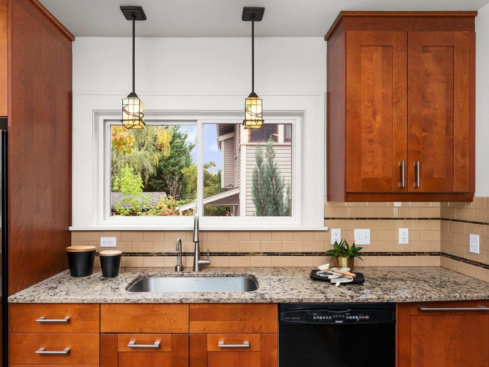 Modern kitchen with wooden cabinets, granite countertop, and stainless steel sink under a large window. Black dishwasher and two pendant lights hanging above. Outdoor scene visible through the window, featuring trees and a neighboring house.