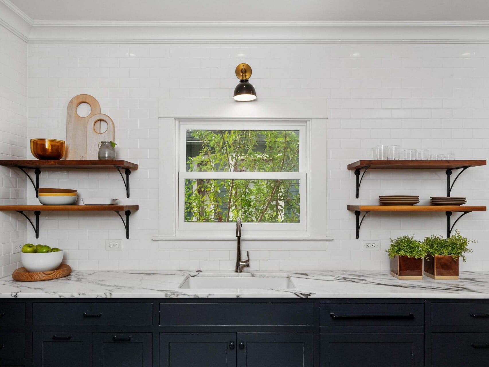 A modern kitchen in Portland, Oregon boasts a black and white color scheme with a marble countertop and dark cabinets. Wooden shelves on either side display dishes and decor items. A central window invites natural light, framed by a single wall light above.