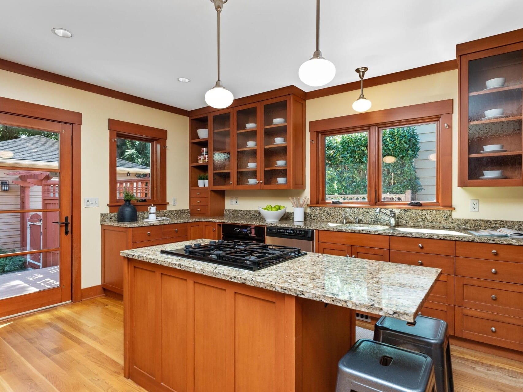 A cozy Portland, Oregon kitchen with wooden cabinets and granite countertops features a central island with a built-in stove. Pendant lights hang overhead, while a window and glass door reveal a charming garden view outside.