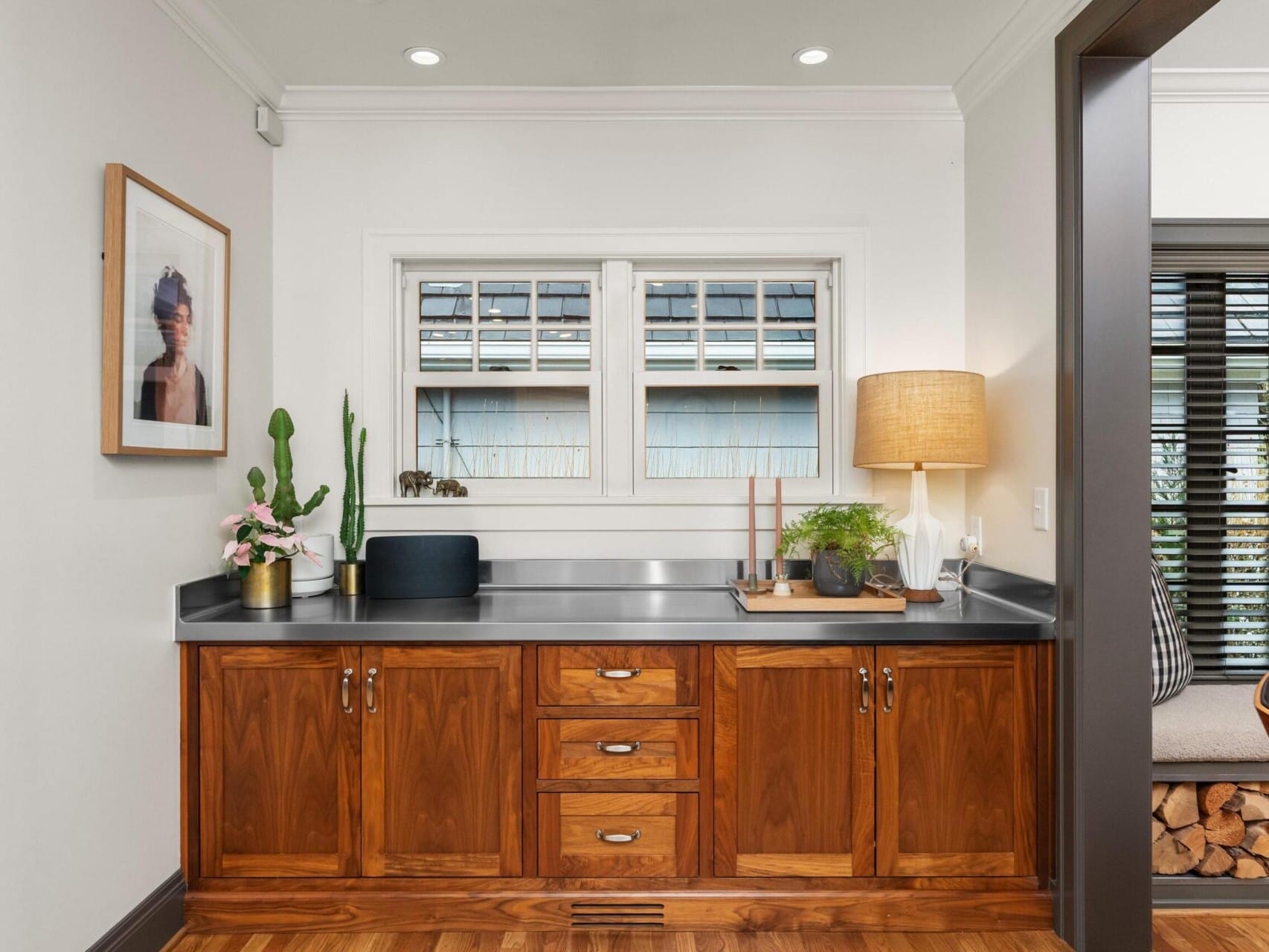 A modern home office with a wooden desk featuring cabinets and drawers is the perfect setting for a Portland Realtor. A lamp, potted plants, and decorative items adorn the desk, while a framed picture hangs on the wall. Light pours in through the window, enhancing this cozy Portland Oregon real estate gem.