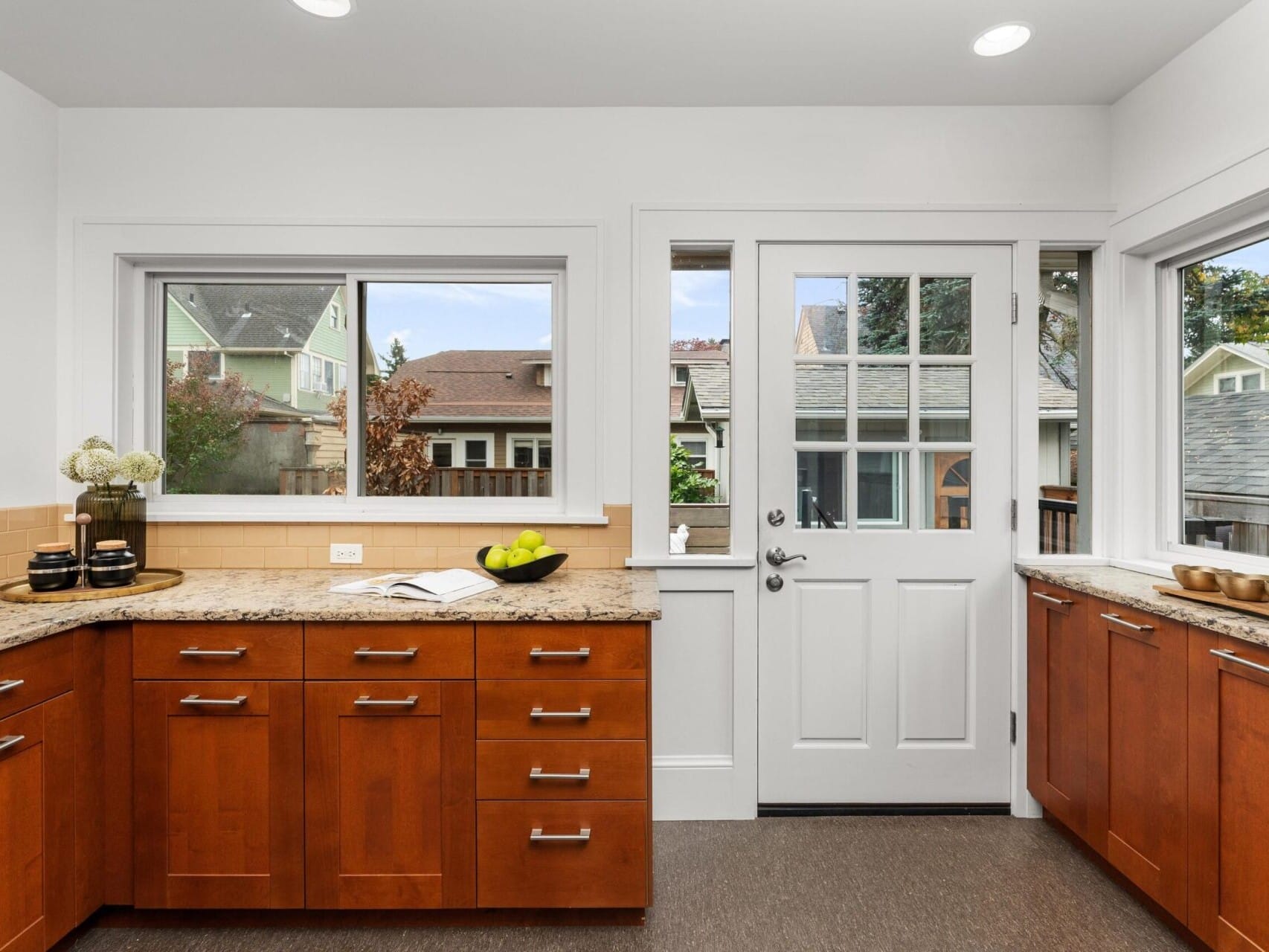 A modern kitchen with wooden cabinets, granite countertops, and a large window. A white door with glass panels opens to the outside. A bowl of green apples and some potted plants decorate the space.