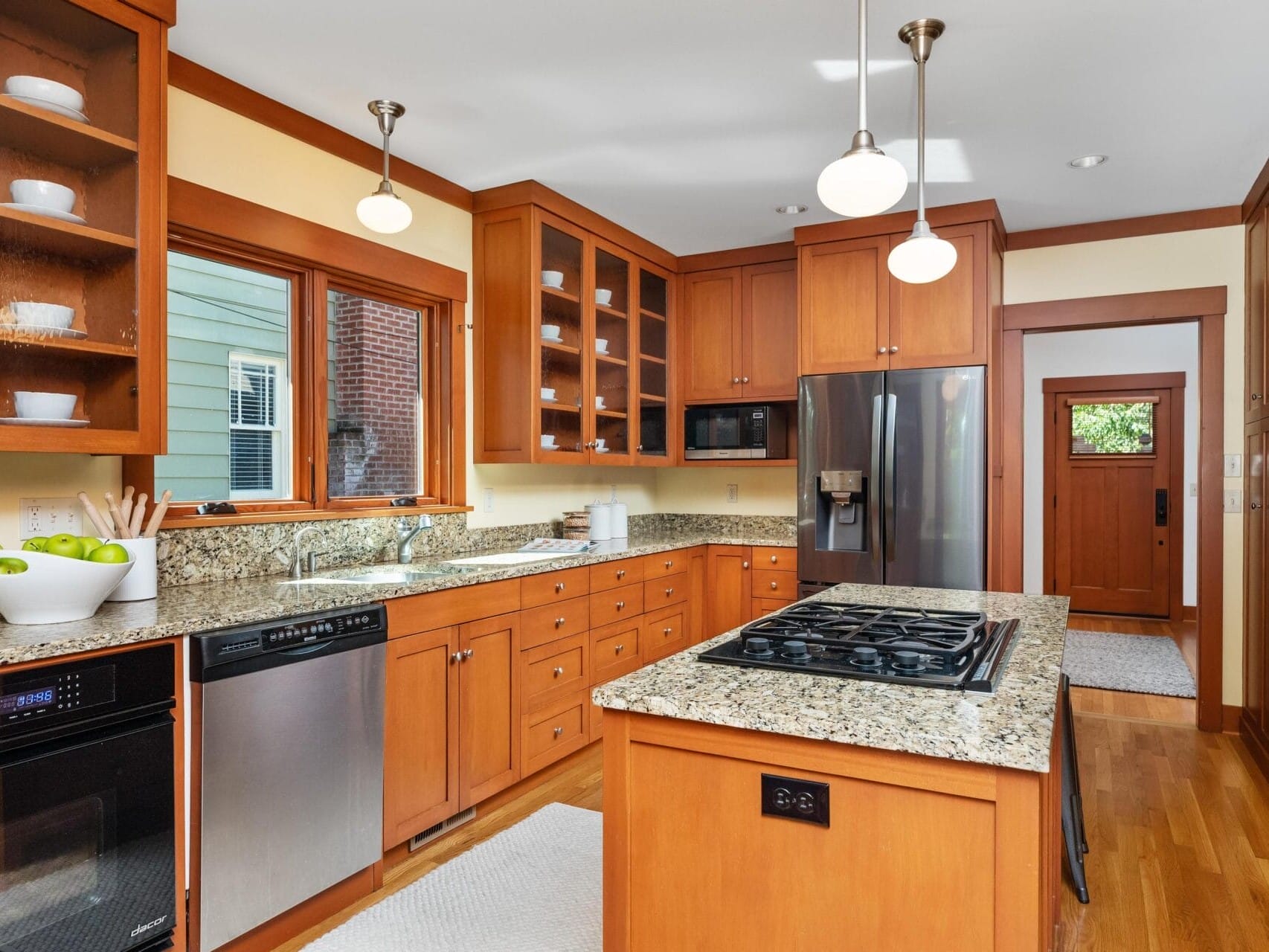 A modern kitchen in Portland, Oregon features wooden cabinets, granite countertops, and stainless steel appliances. Pendant lights illuminate a gas stove on the island, while a window overlooks the sink area. A bowl of green apples adds a fresh touch to the counter.