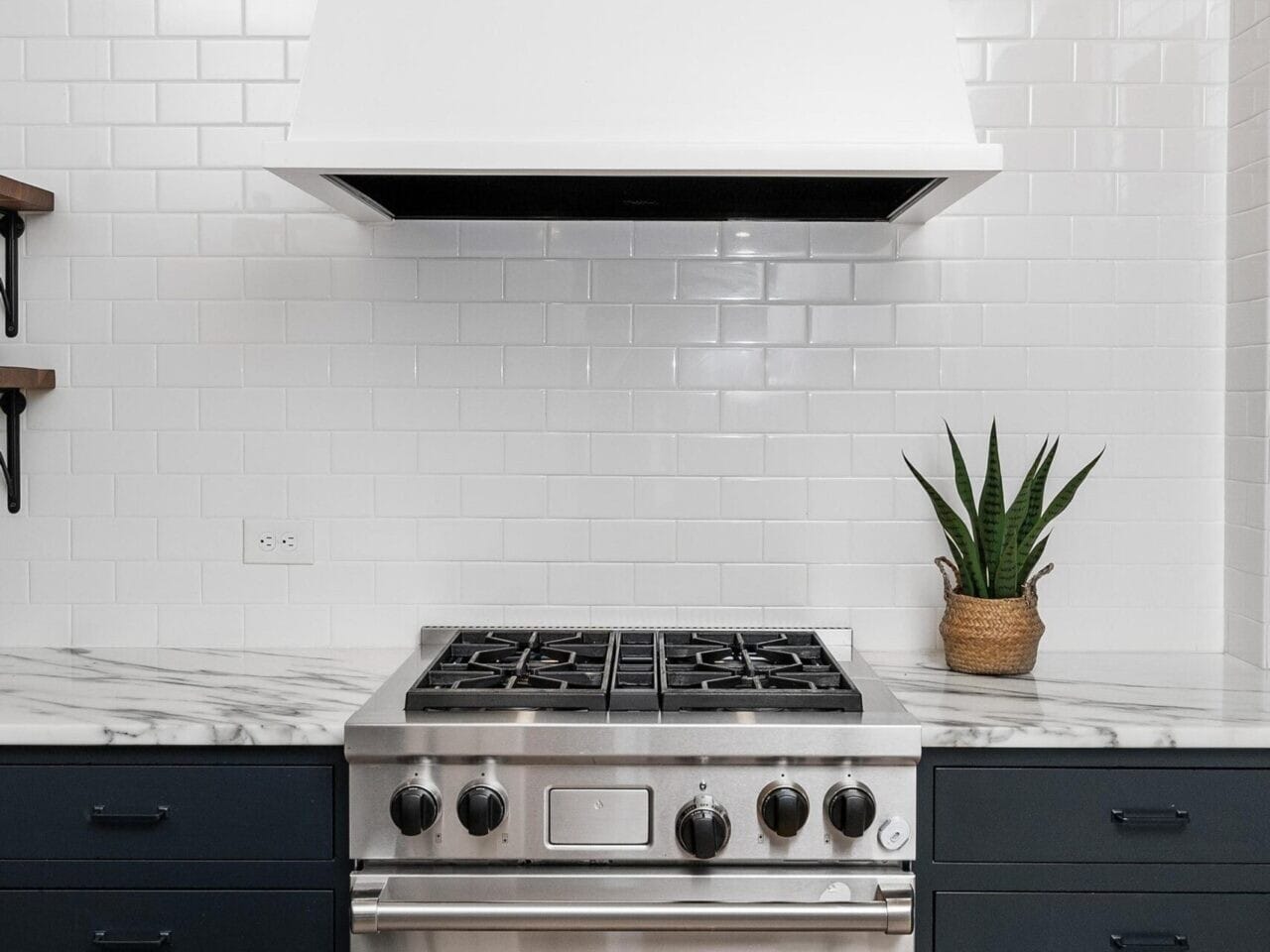 A modern Portland, Oregon kitchen features a stainless steel gas stove and overhead white range hood set against a white tiled backsplash. The sleek countertop is marble with navy blue cabinets below, complemented by a potted plant on the right side.