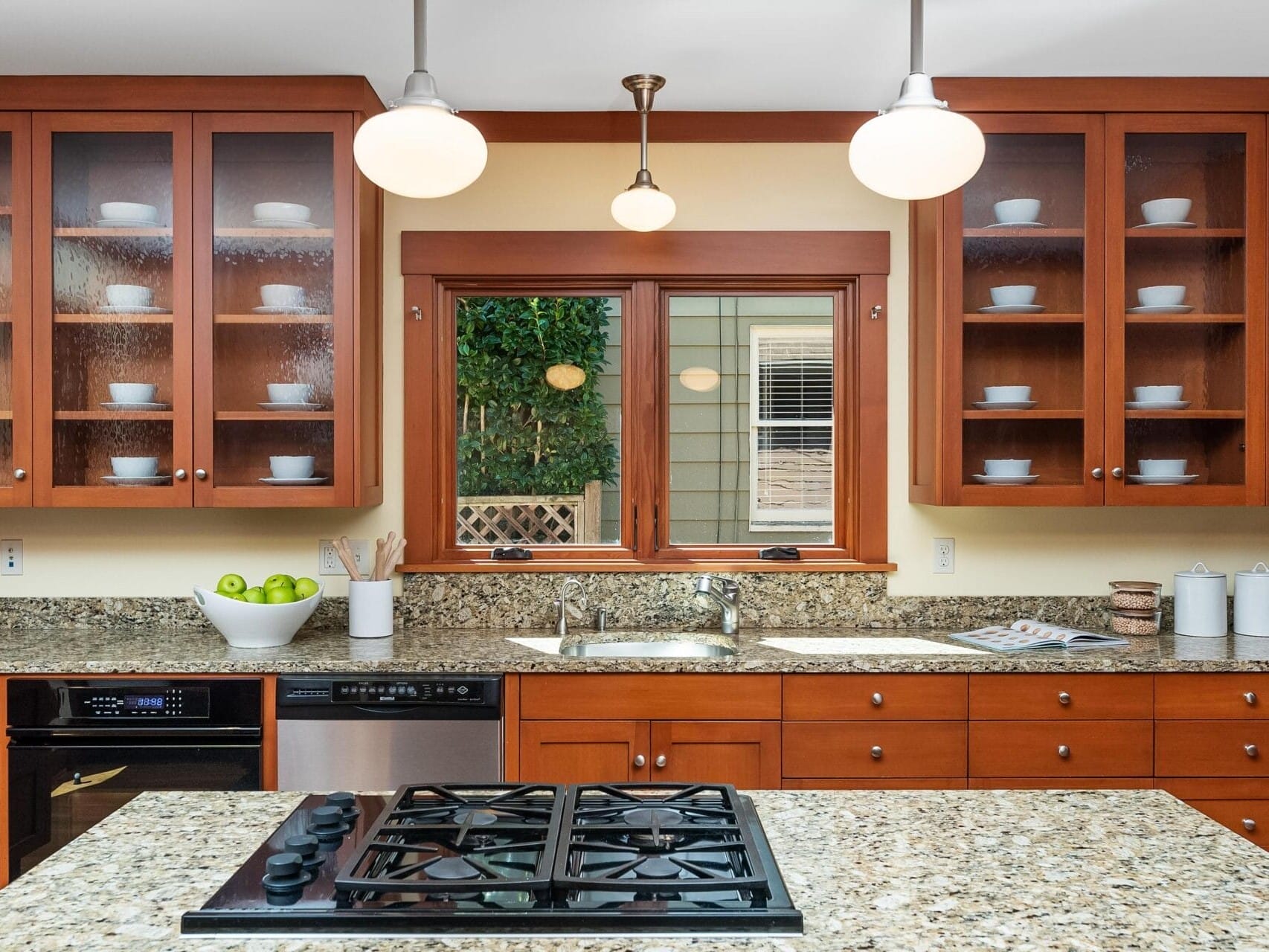 A kitchen in Portland, Oregon, with wooden cabinets and a marble countertop features a built-in stove. The cabinets display white dishes, while a bowl of green apples sits on the counter. Pendant lights hang from the ceiling, and a window overlooks lush greenery outside.