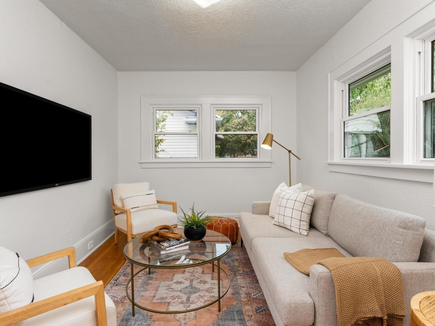 A cozy living room features a light gray sofa with a brown throw, a round glass coffee table, two armchairs, a floor lamp, and a wall-mounted TV. Three windows let in natural light, and a patterned rug adds warmth to the hardwood floor.