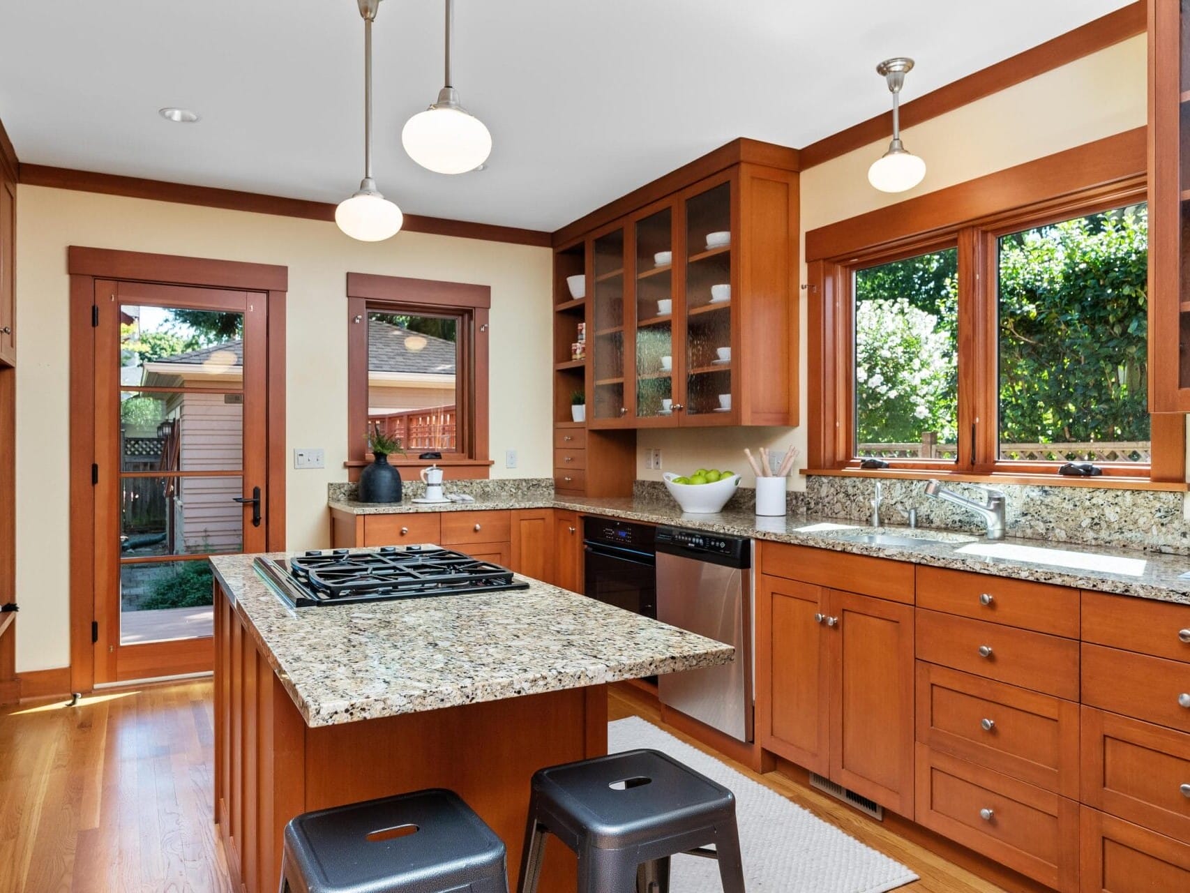 A modern Portland, Oregon kitchen boasts wooden cabinets, granite countertops, and a central island with a gas stove. Two bar stools are tucked under the island. Large windows offer natural light, while a door leads to the garden. Pendant lights gracefully hang from the ceiling.