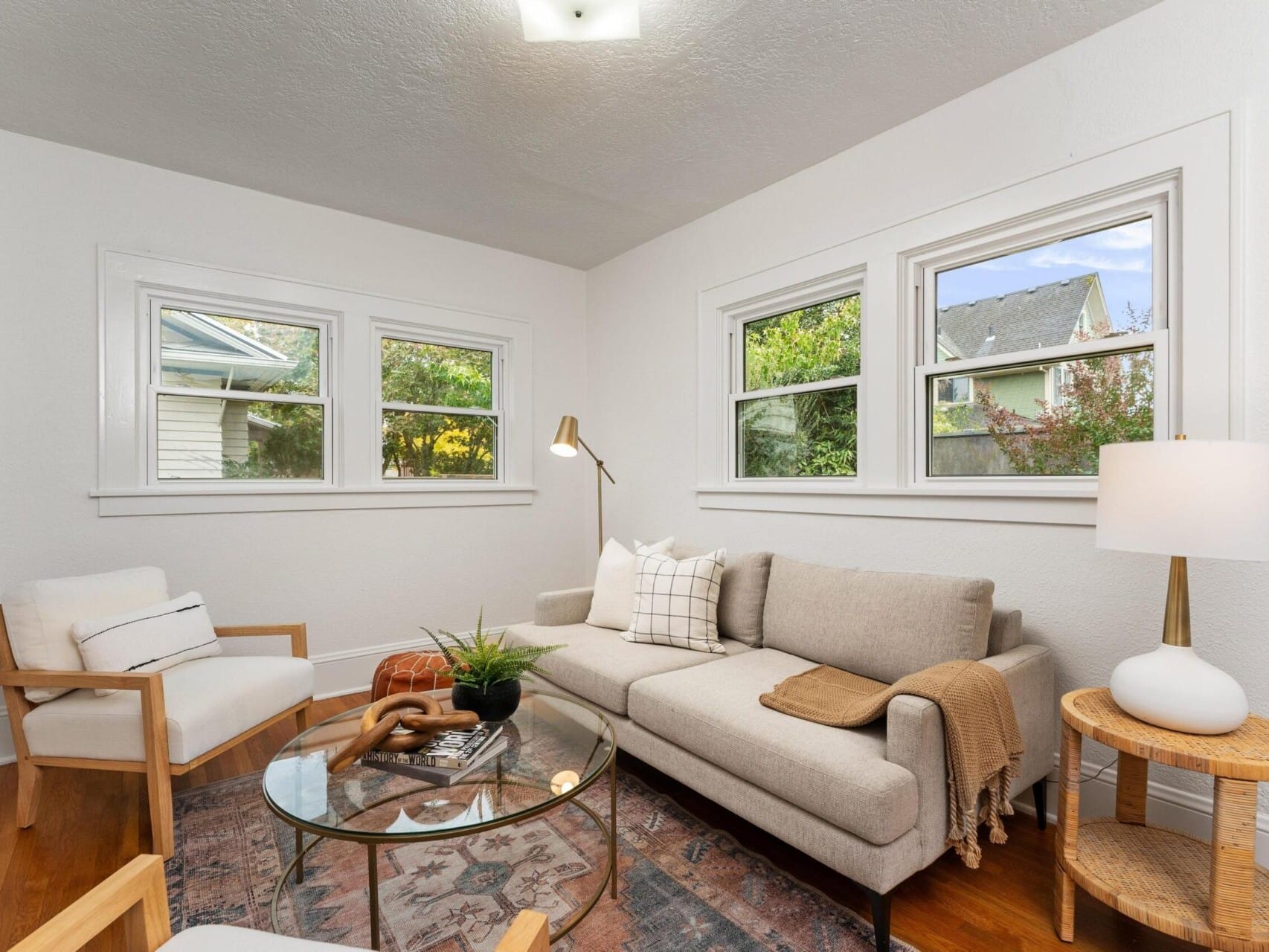 A cozy living room with a beige sofa, armchair, and a glass coffee table. A floor lamp and table lamp provide light. The walls are white, and three windows offer a view of greenery outside. A patterned rug covers the wooden floor.