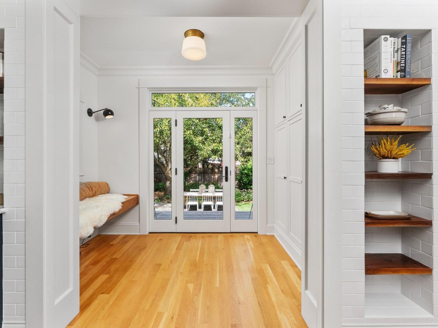 Bright, minimalist room in Portland, Oregon, with wooden flooring, white walls, and open double doors leading to a garden. A bench with a cushion is on the left, and built-in shelves on the right hold books and decor. A soft light fixture hangs from the ceiling.