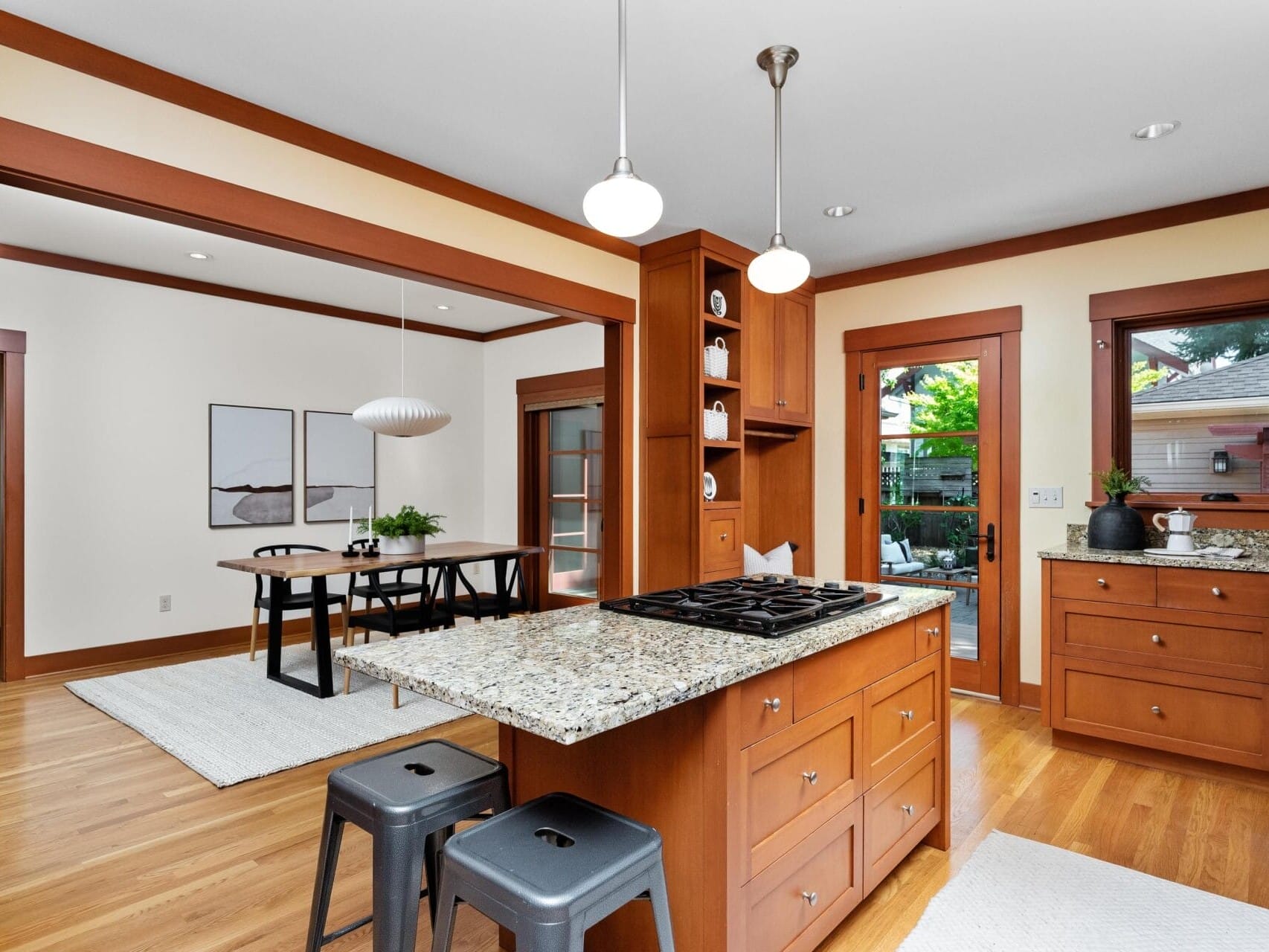 A modern kitchen and dining area in Portland, Oregon, features wooden cabinets, granite countertops, and a kitchen island with stools. Pendant lights hang from the ceiling. A dining table with chairs is visible in the adjoining room. Large windows and a door lead outside.