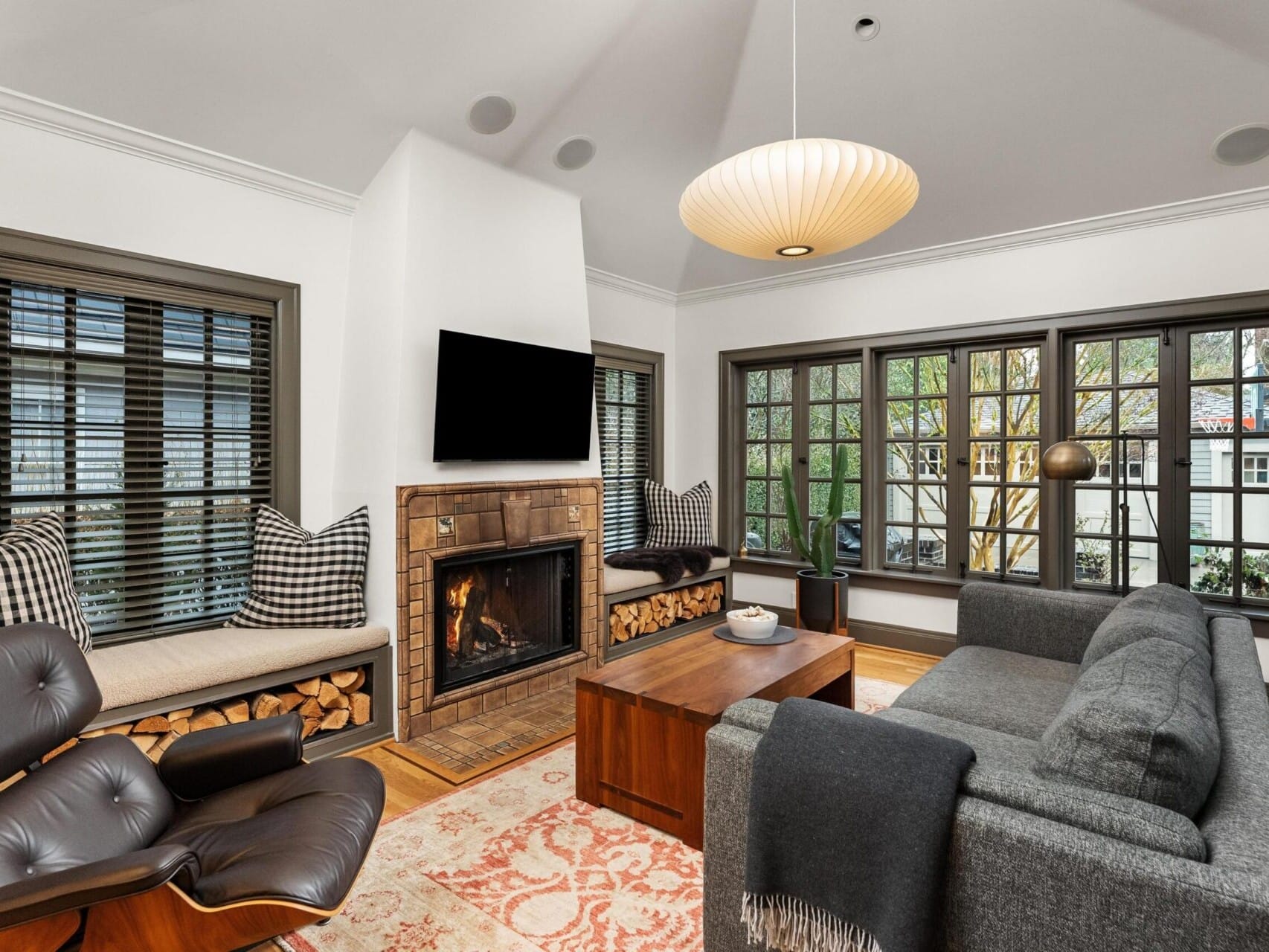 A cozy living room in Portland, Oregon real estate features a modern fireplace surrounded by stacked wood. A dark gray sofa, armchair, and wooden coffee table create a welcoming ambiance. Large grid-patterned windows fill the space with natural light, while a stylish pendant light hangs overhead.