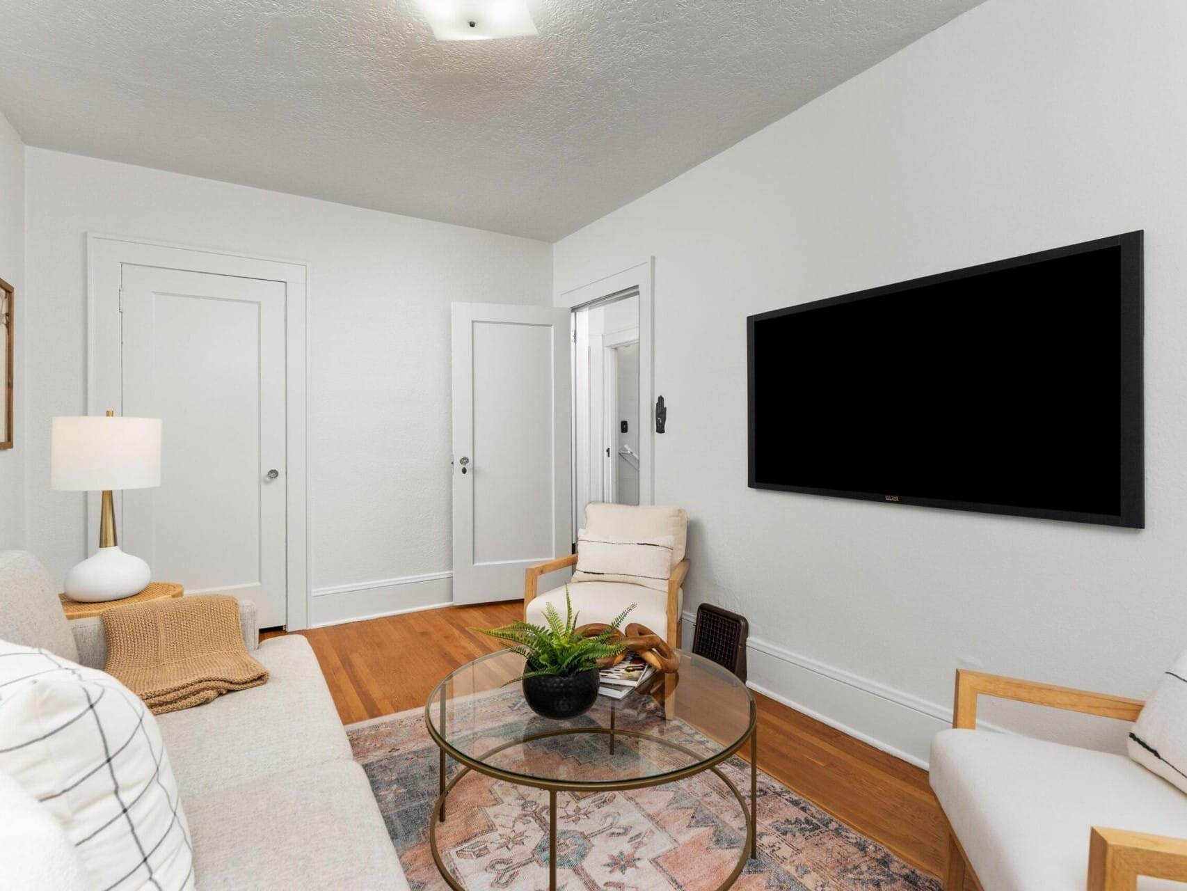 A cozy living room with white walls, featuring a cushioned sofa, armchairs, a glass coffee table with a plant, and a TV mounted on the wall. A lamp provides soft lighting, and a patterned rug adds warmth to the wooden floor.