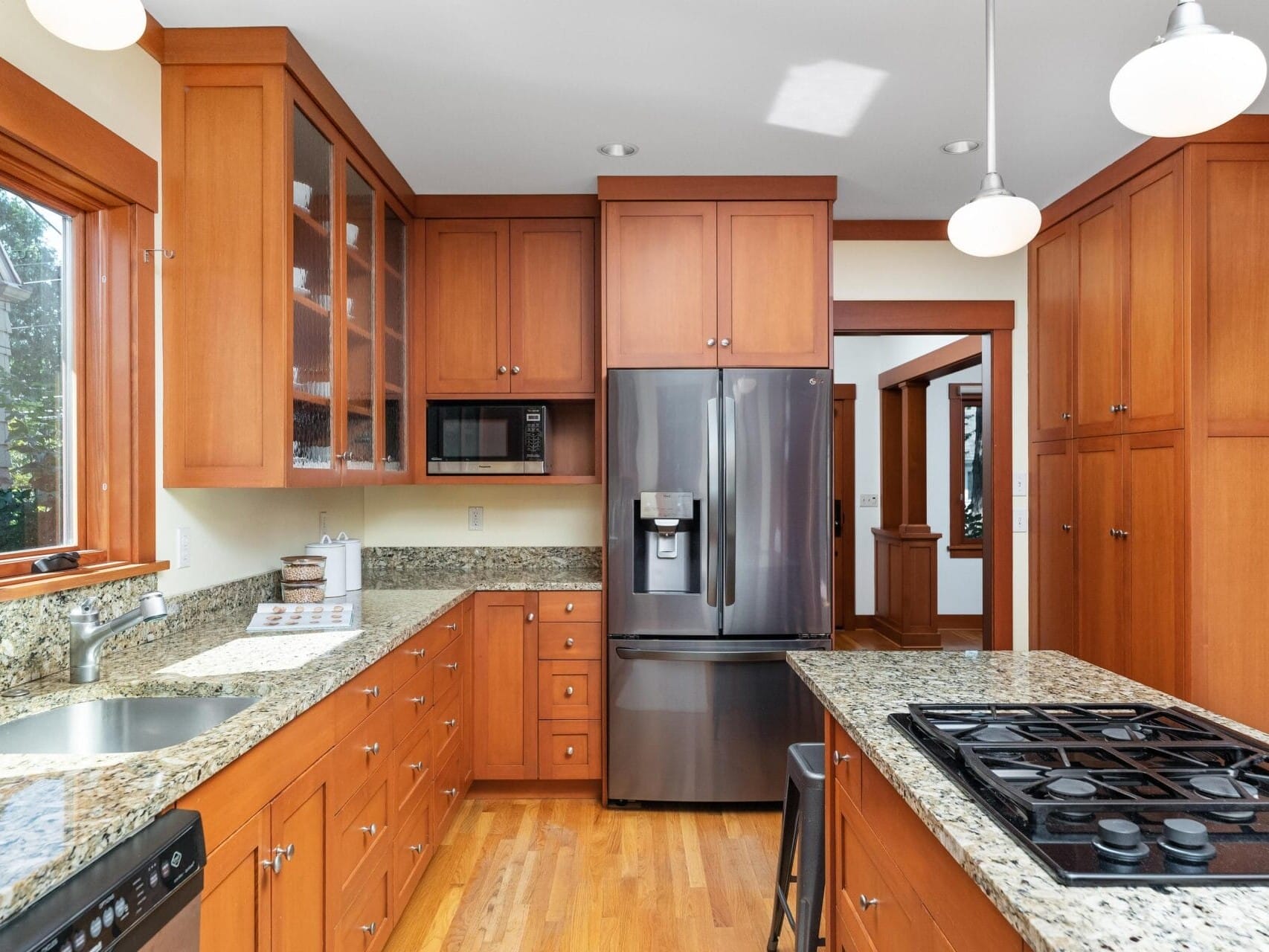 A modern kitchen in Portland, Oregon, with wooden cabinets and granite countertops. It features a stainless steel refrigerator, oven, and gas stovetop on a central island. Pendant lights hang from the ceiling, and large windows let in natural light.