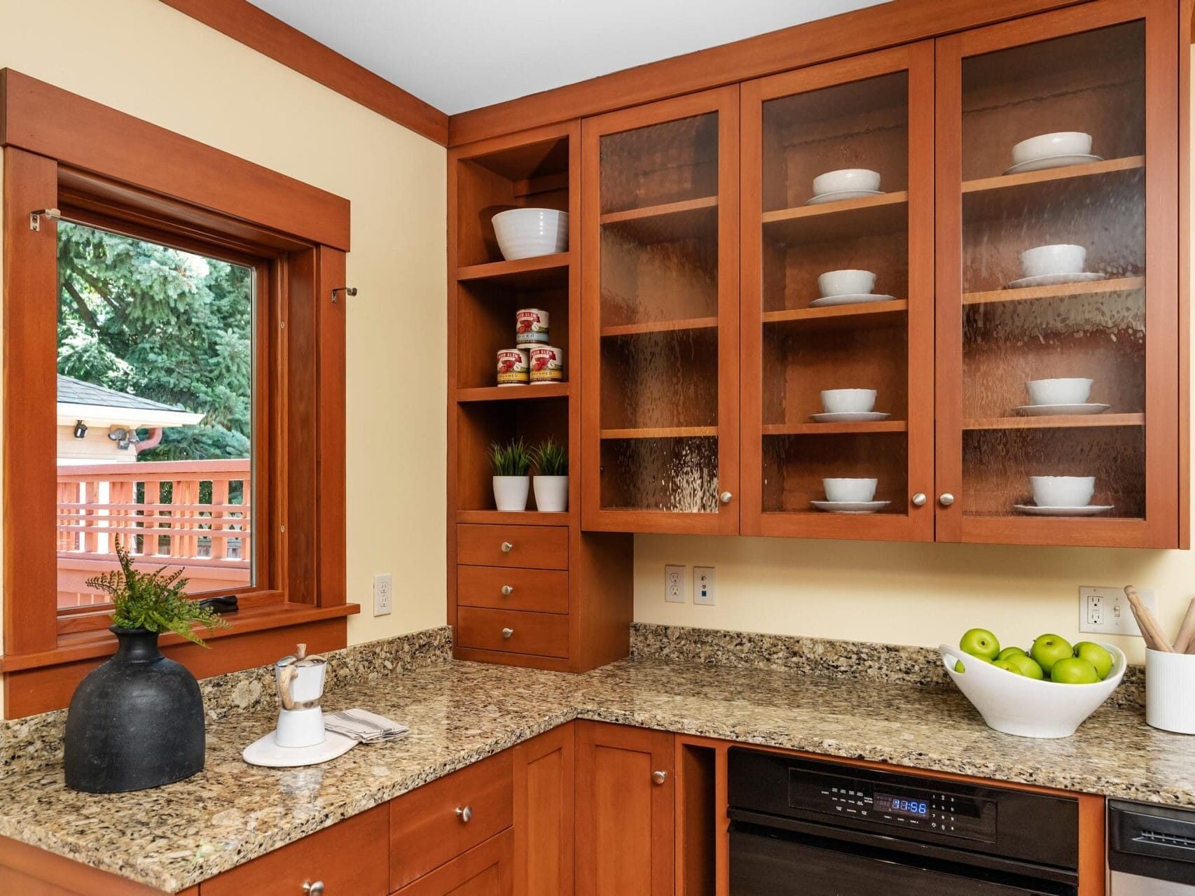 A cozy Portland, Oregon kitchen corner features wooden cabinets with glass doors displaying white dishes. The countertops host a potted plant, black vase, and a bowl of green apples. Through the window, a deck overlooks lush trees, enhancing the warm and inviting atmosphere.