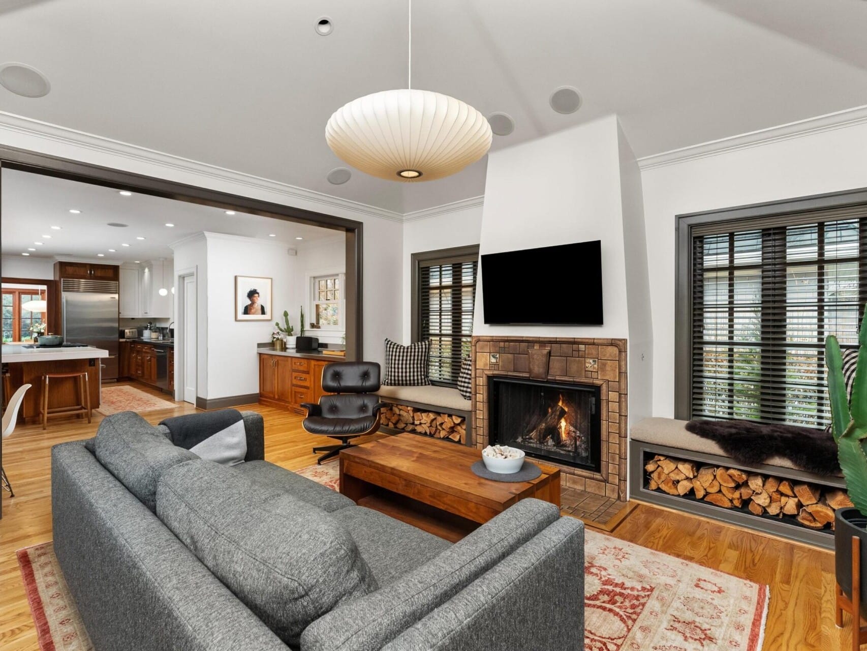 A cozy living room with a gray sofa, wooden coffee table, and a fireplace. Firewood is stacked under the window seats, and a flat-screen TV is mounted above the fireplace. Perfect for entertaining, it opens into a modern kitchen with stainless steel appliances, ideal for Portland Oregon real estate seekers.