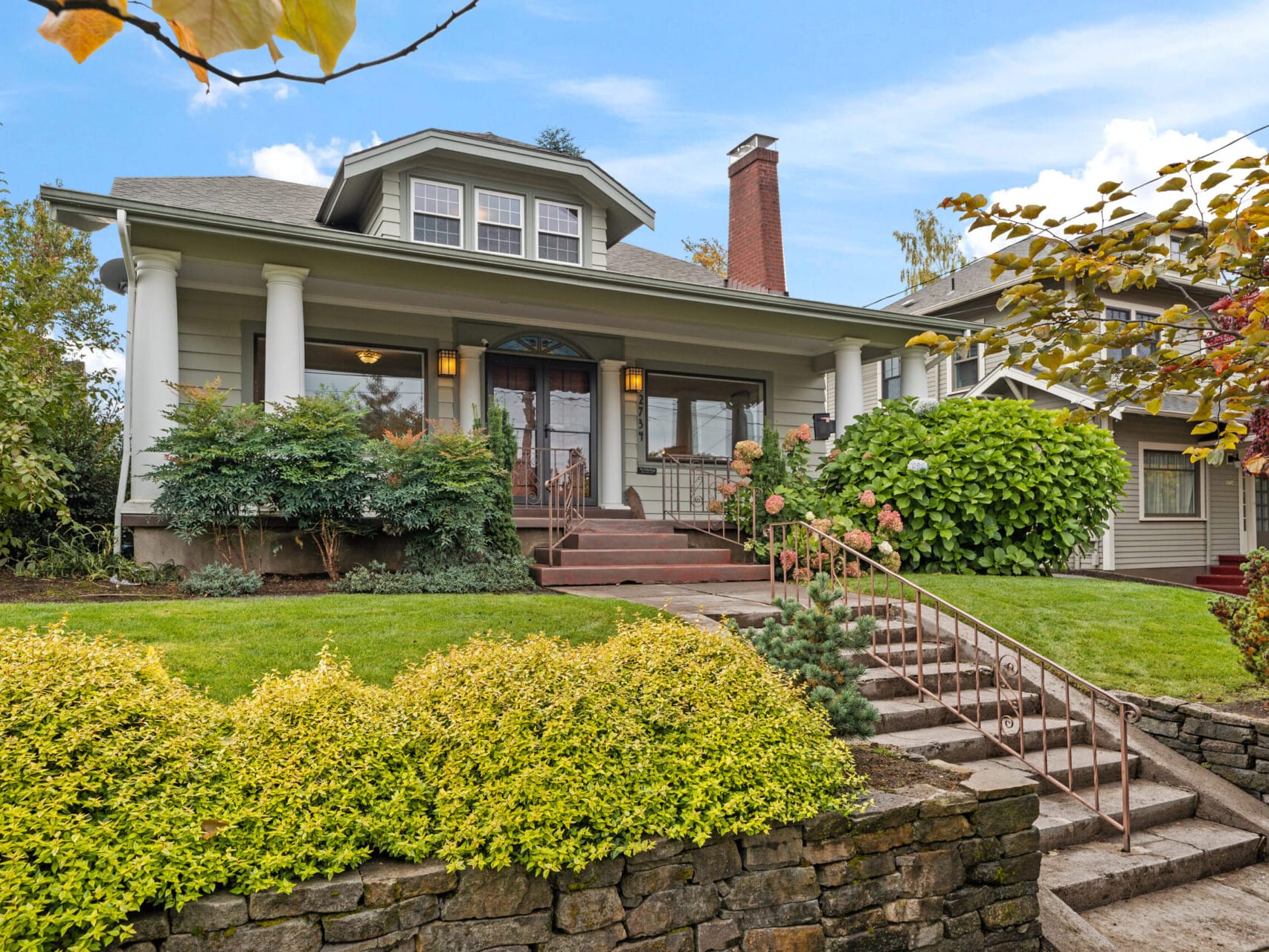 Discover this charming two-story house in Portland, where a brick chimney complements the large front porch, with stairs leading up through a well-manicured garden. Surrounded by trees and shrubs under a bright blue sky, it's every Portland Oregon real estate enthusiast's dream.