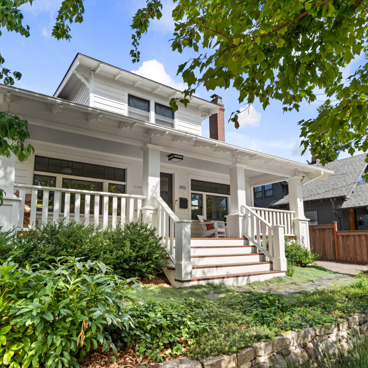 A charming white two-story house with a spacious porch surrounded by lush greenery and trees offers the quintessential Portland, Oregon, real estate charm. The blue sky and sunlight create a welcoming atmosphere, with steps leading up to the porch accented by white railings and cozy seating.