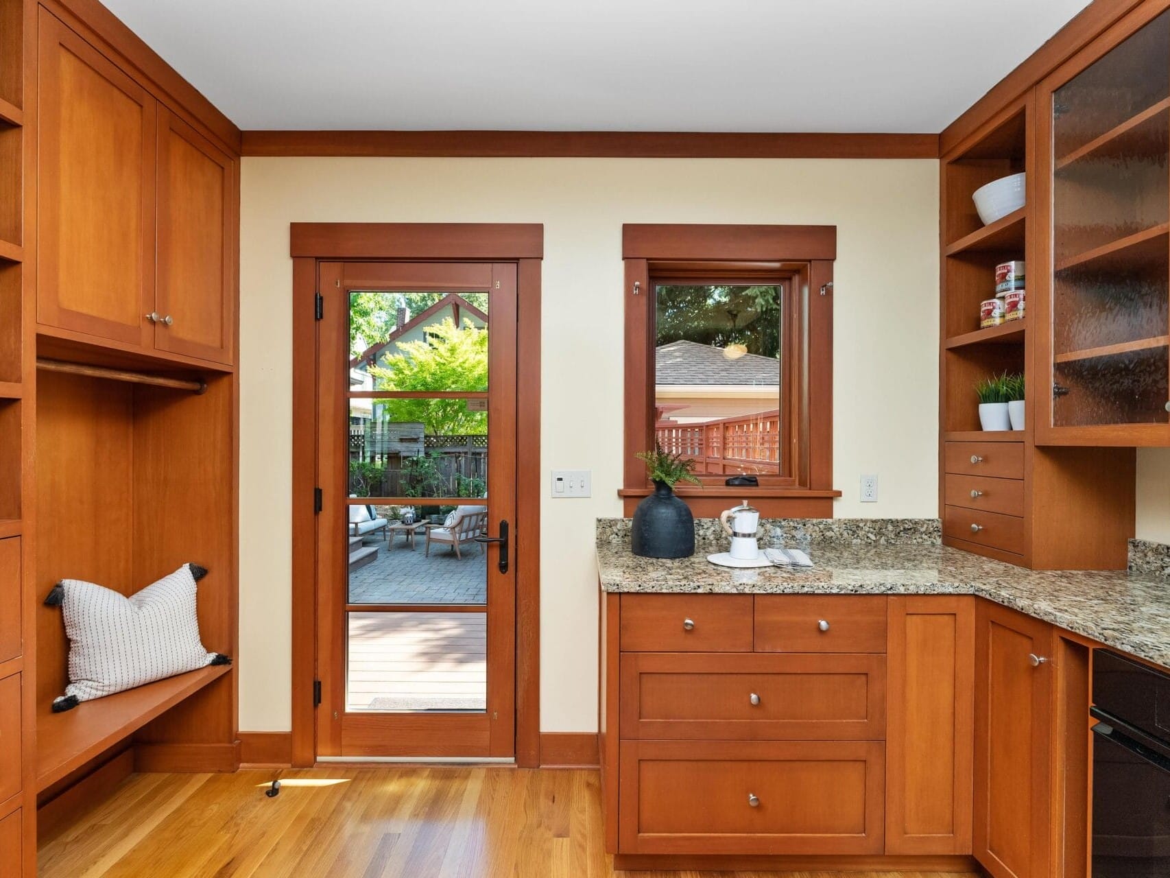 A warm, wooden kitchen with granite countertops in Portland, Oregon features a glass door leading to an outside area. Cabinets brim with dishes while a plant adorns the countertop. Natural light flows in, highlighting the wooden floors and built-in bench.