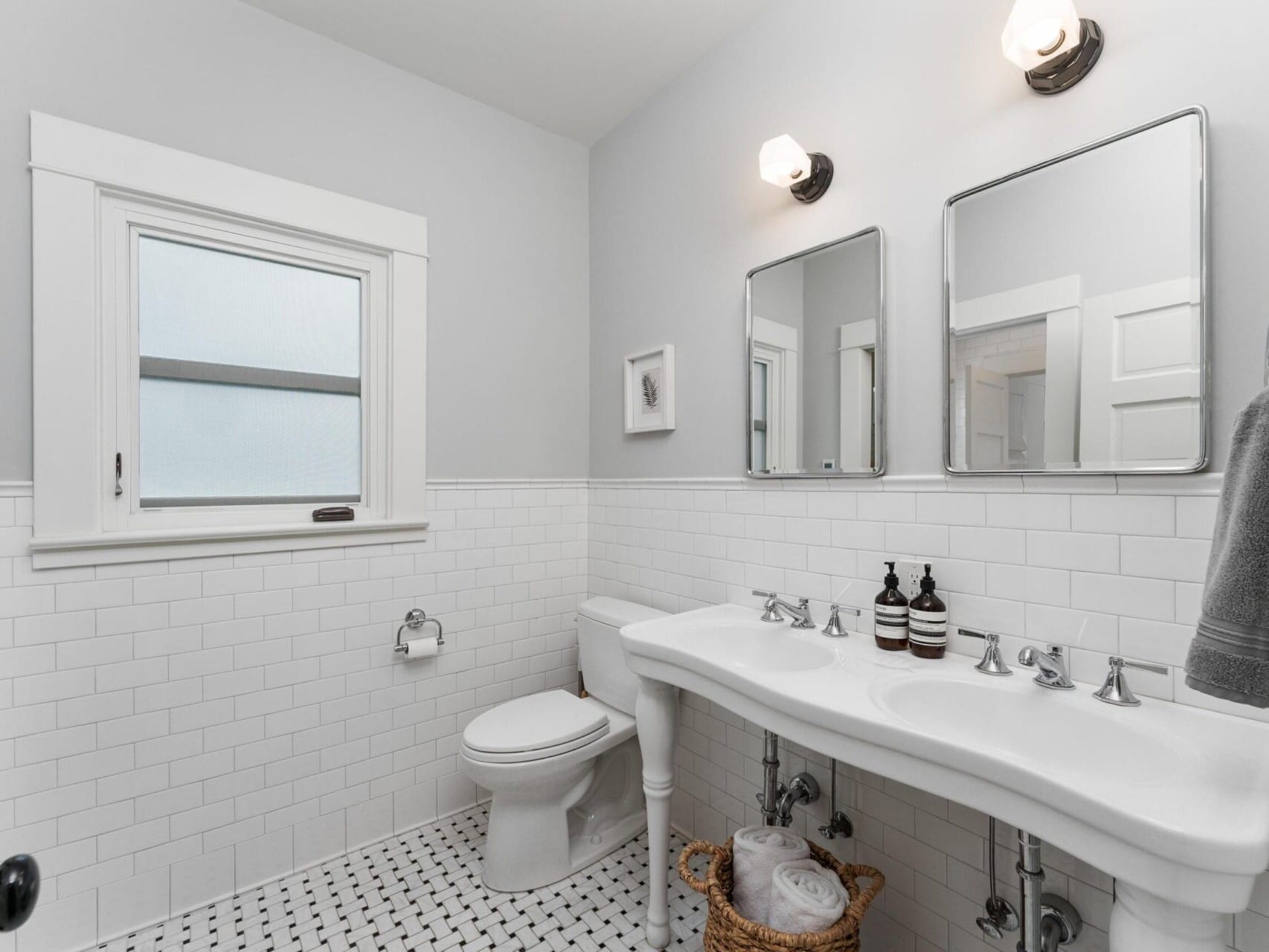 A bright bathroom in a Portland, Oregon style features white walls and subway tiles. It includes a double sink with two mirrors, a toilet, and a window. A towel hangs on the wall, toiletries rest on the sink, and a basket with towels sits on the floor.